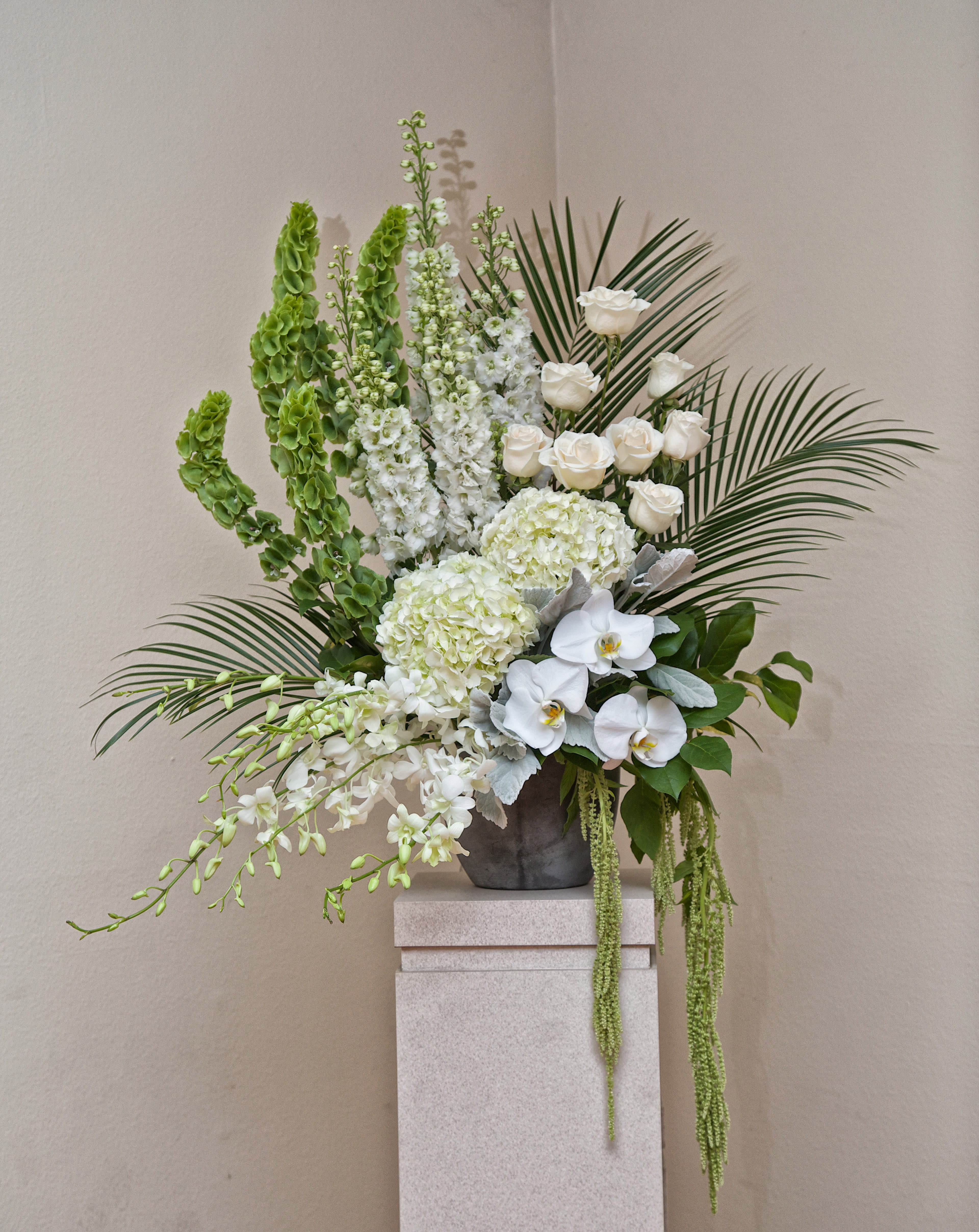 Tall white floral arrangement with orchids, roses, and palm fronds in a gray vase