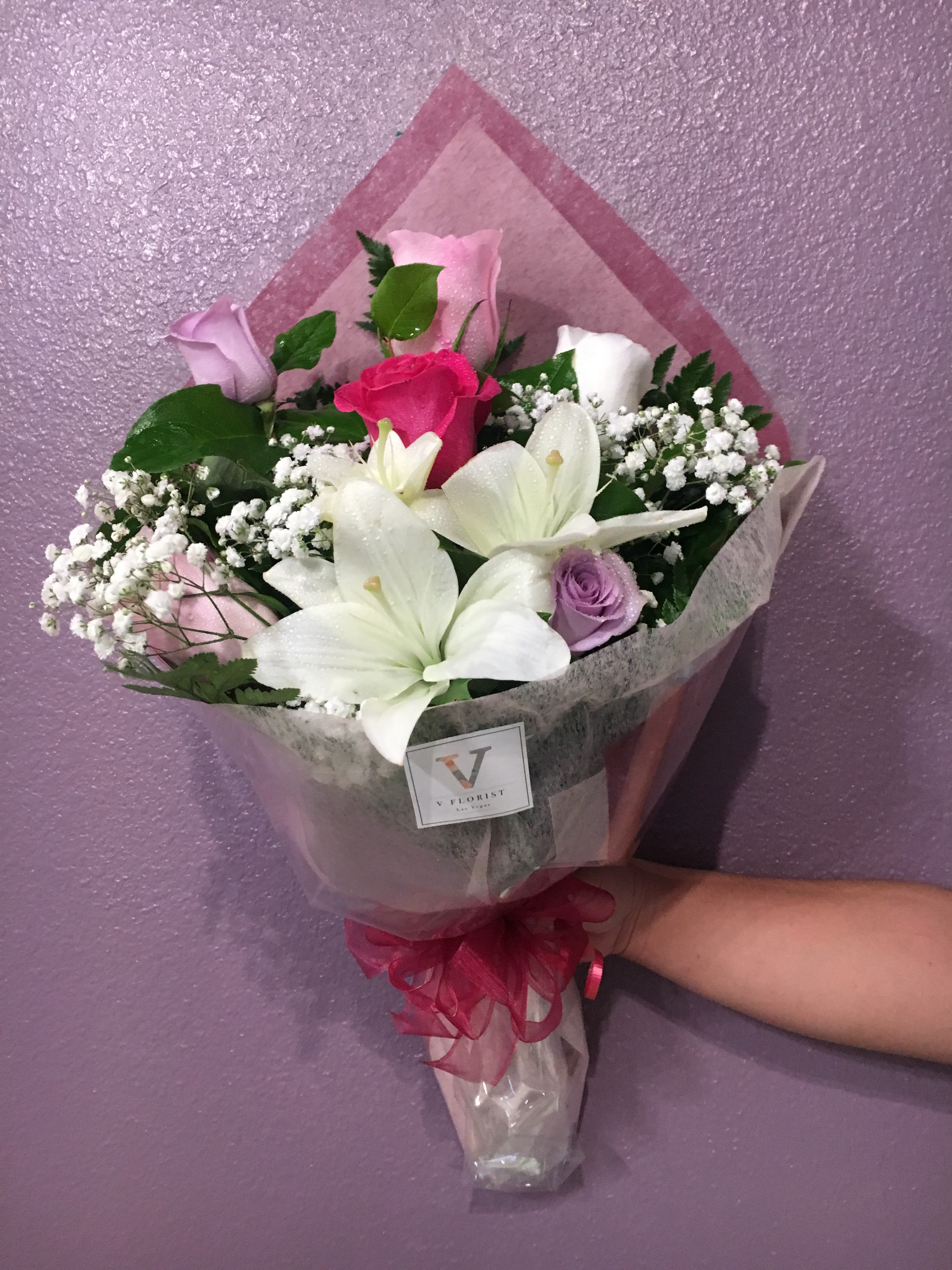 Wrapped bouquet of white lilies and mixed pink and purple roses held against a purple wall