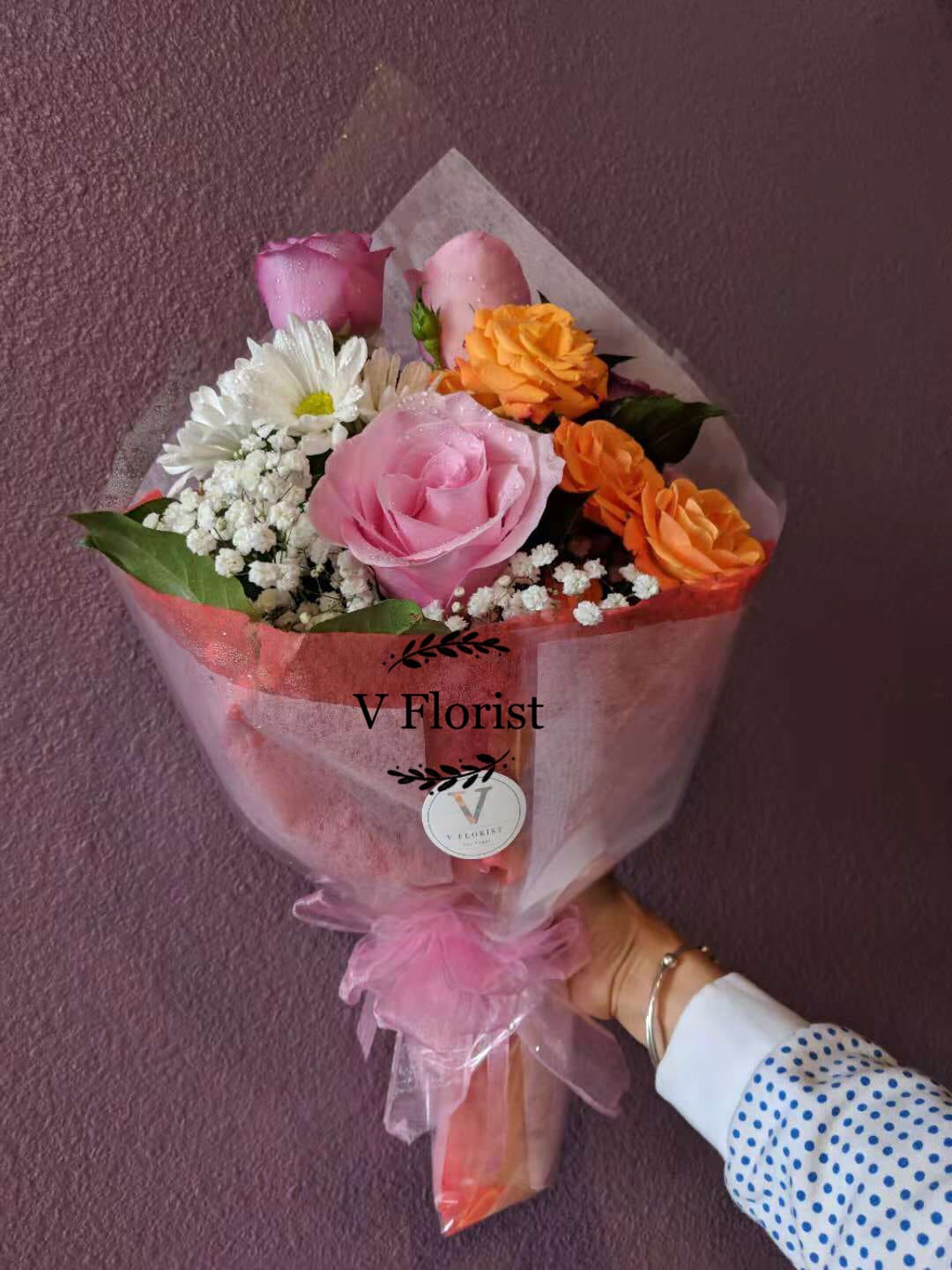 Hand-tied bouquet of pink roses, orange roses, white daisies and baby’s breath wrapped in pink paper.