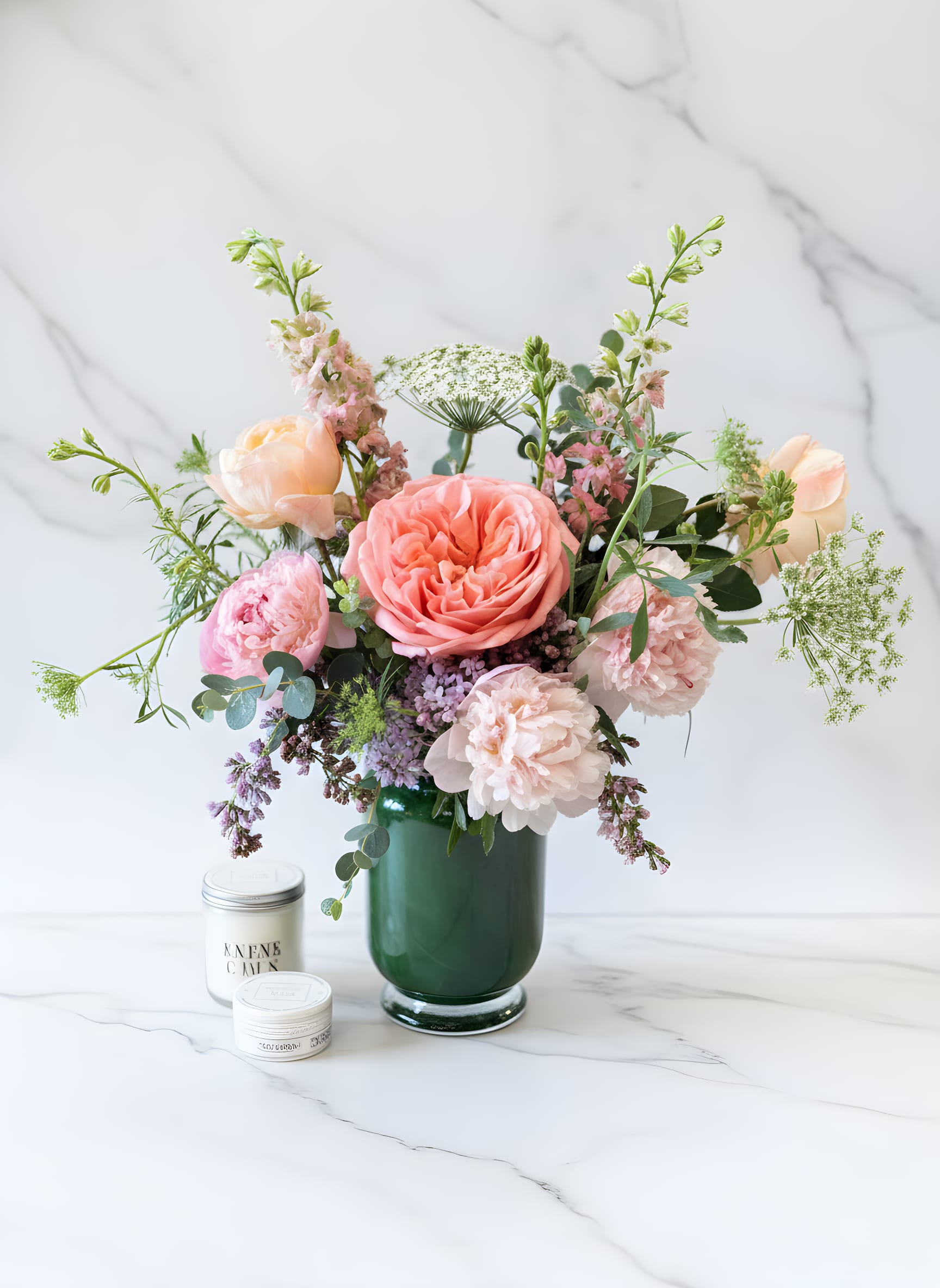 Pastel pink and peach flower arrangement in a dark green glass vase with two small white candle tins.