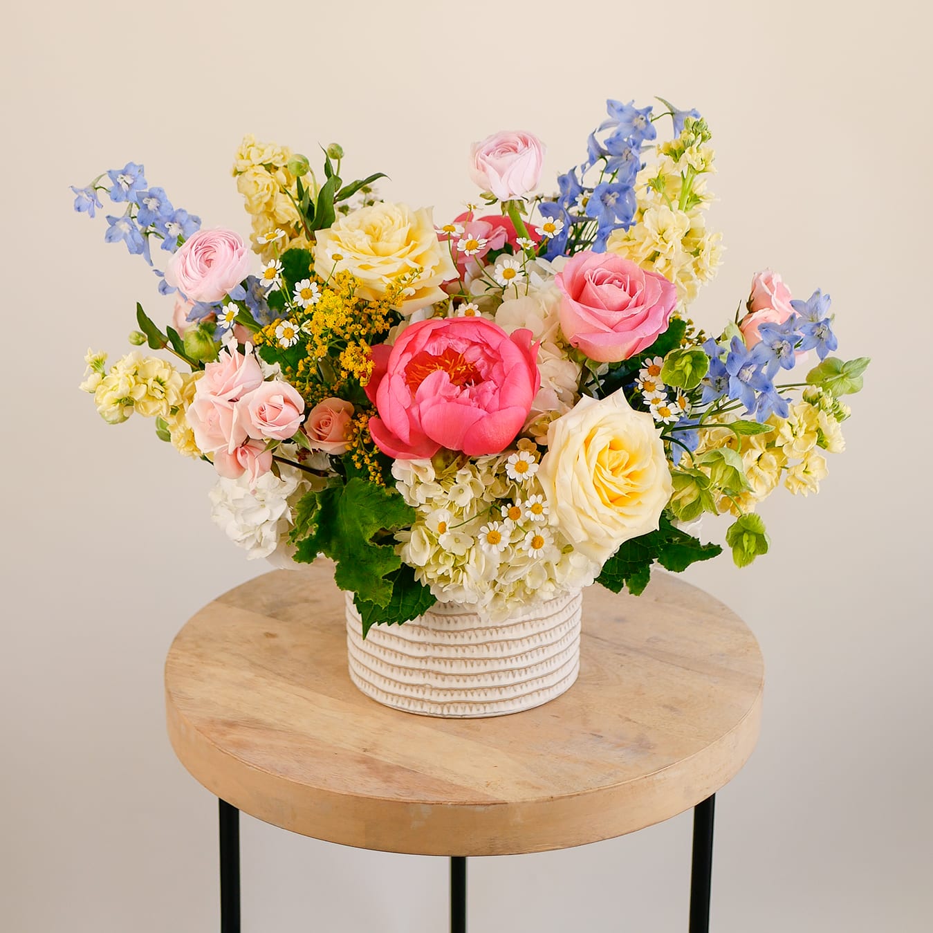 Summer Spotlight - Bursting with color and charm, "English Garden" is a garden-inspired delight featuring vibrant coral peonies, delicate ranunculus, soft roses, pale blue delphinium, yellow stock, and playful chamomile. Artfully arranged in a textured ceramic container, this fresh and whimsical design brings the feeling of a blooming meadow into any space. Ideal for spring celebrations, birthdays, or a joyful pick-me-up.