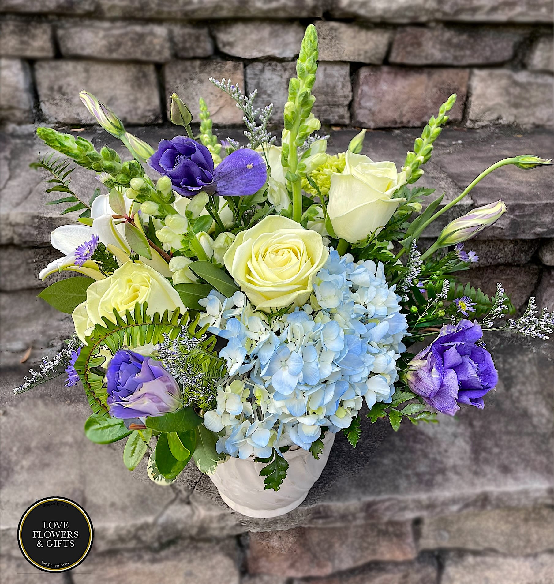 Arrangement of pale yellow roses, blue hydrangeas, and purple blooms in a white vase on stone steps
