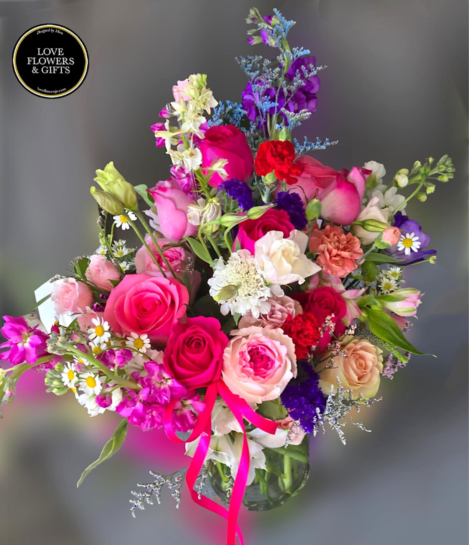 Mixed bouquet of pink roses, carnations, and daisies in a clear glass vase with bright pink ribbon.