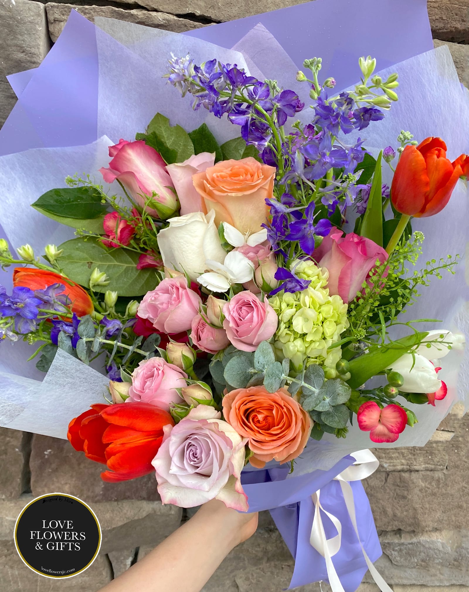 Hand-tied bouquet of mixed roses, tulips, and purple delphinium wrapped in lilac paper.