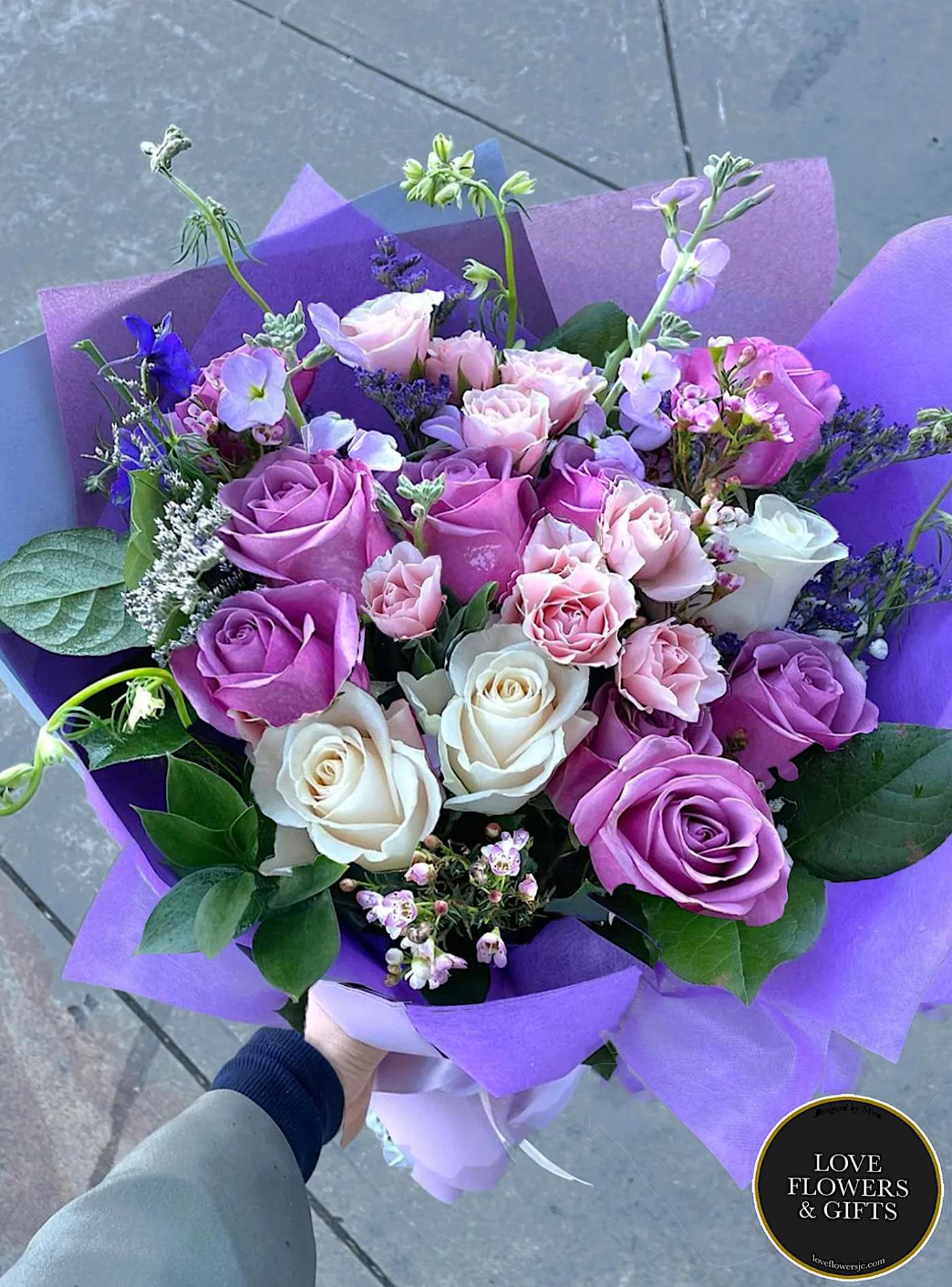 Hand-tied bouquet of lavender, pink, and white roses with accent flowers wrapped in purple paper.