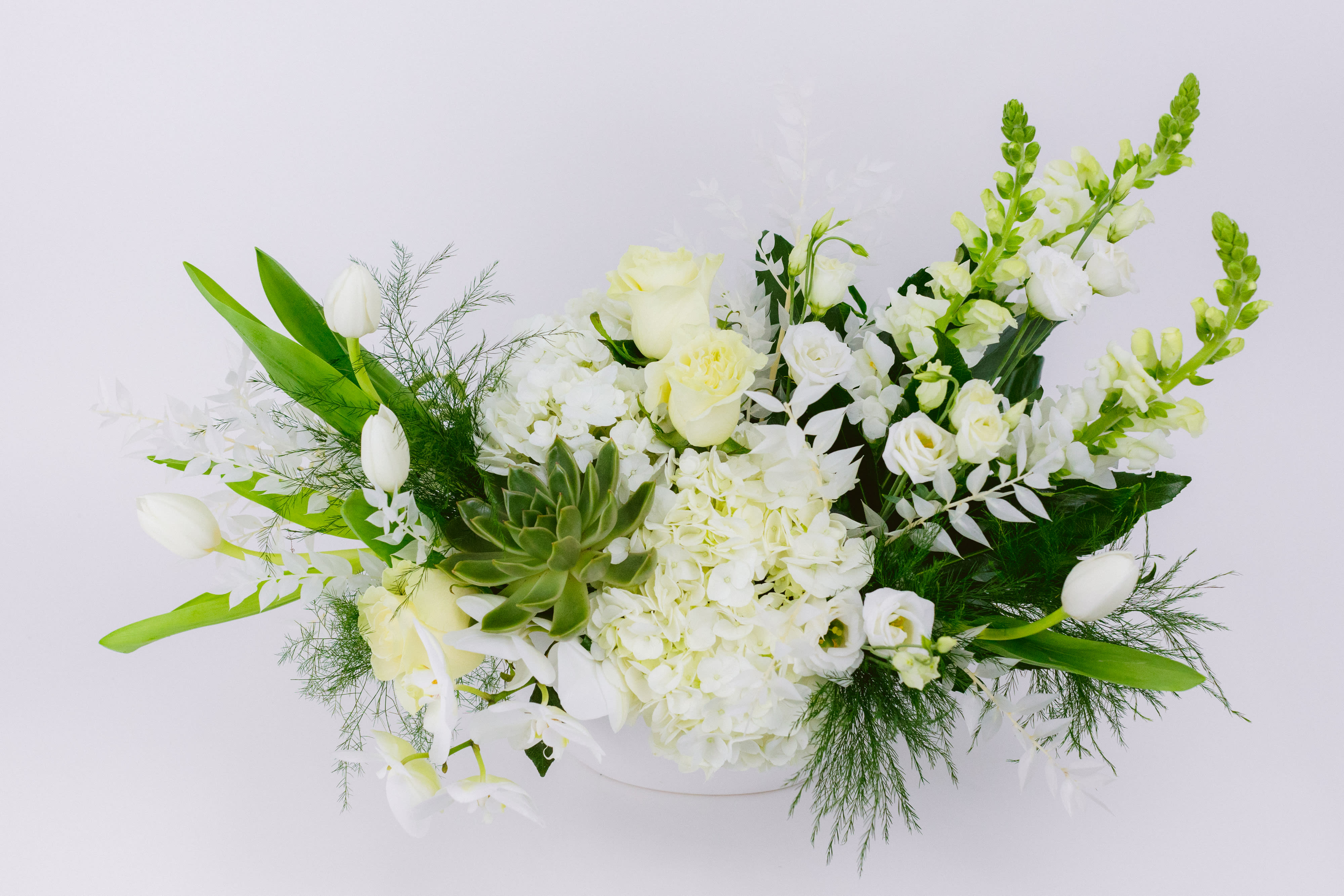 White floral arrangement with tulips, roses, and orchids in a low container