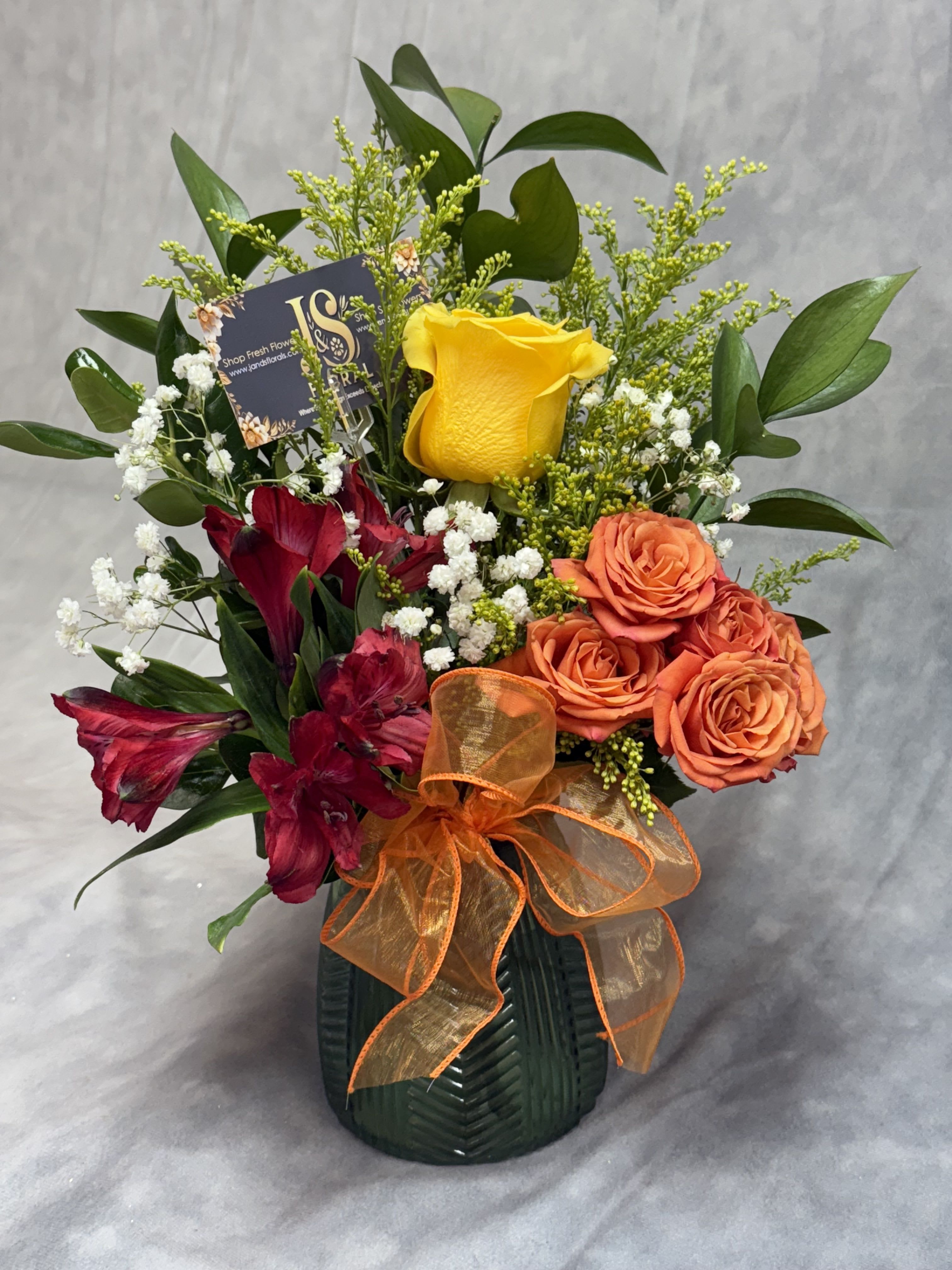 Golden Harvest Vase -  Golden Harvest Vase  A cheerful burst of autumn beauty! This petite arrangement features radiant yellow roses, vibrant orange spray roses, and deep red alstroemeria, accented with lush greenery and seasonal fillers. Designed in a stylish vase, this piece brings a warm and welcoming touch — perfect for brightening any space or gifting a little sunshine.