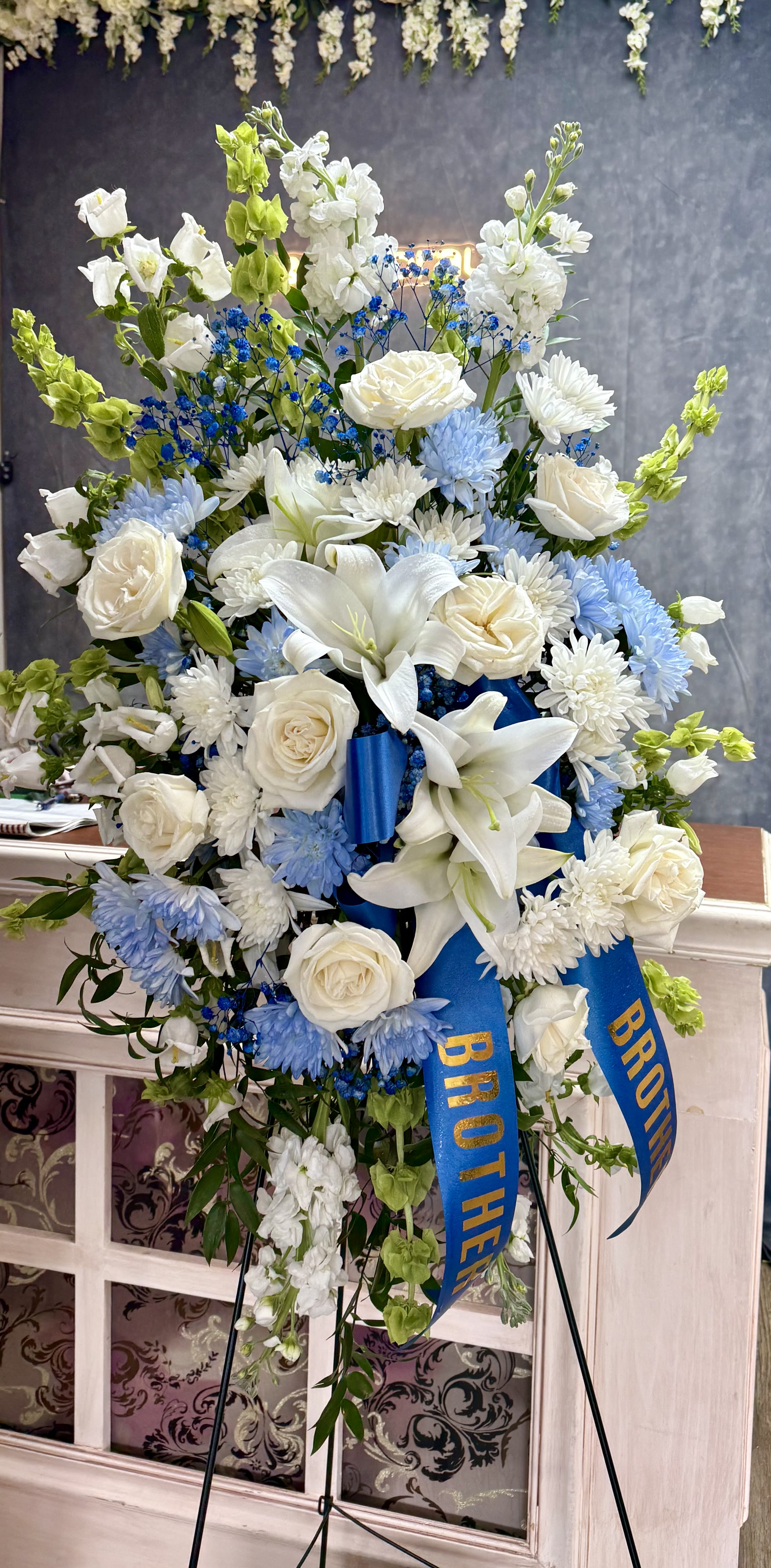 Standing floral spray of white lilies, roses, and blue flowers with a blue ribbon