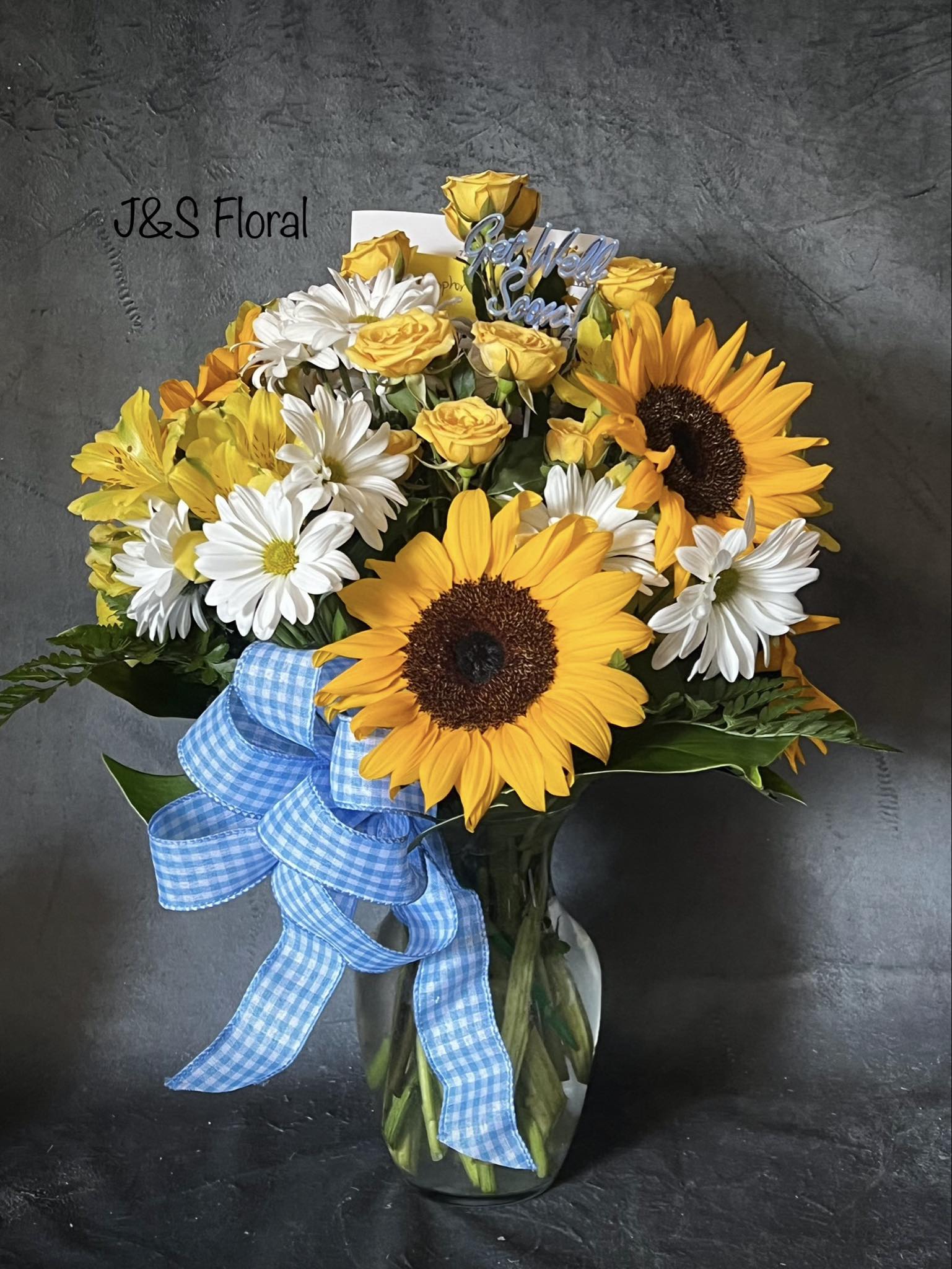 Bouquet of sunflowers, white daisies, and yellow roses in a glass vase with a blue ribbon