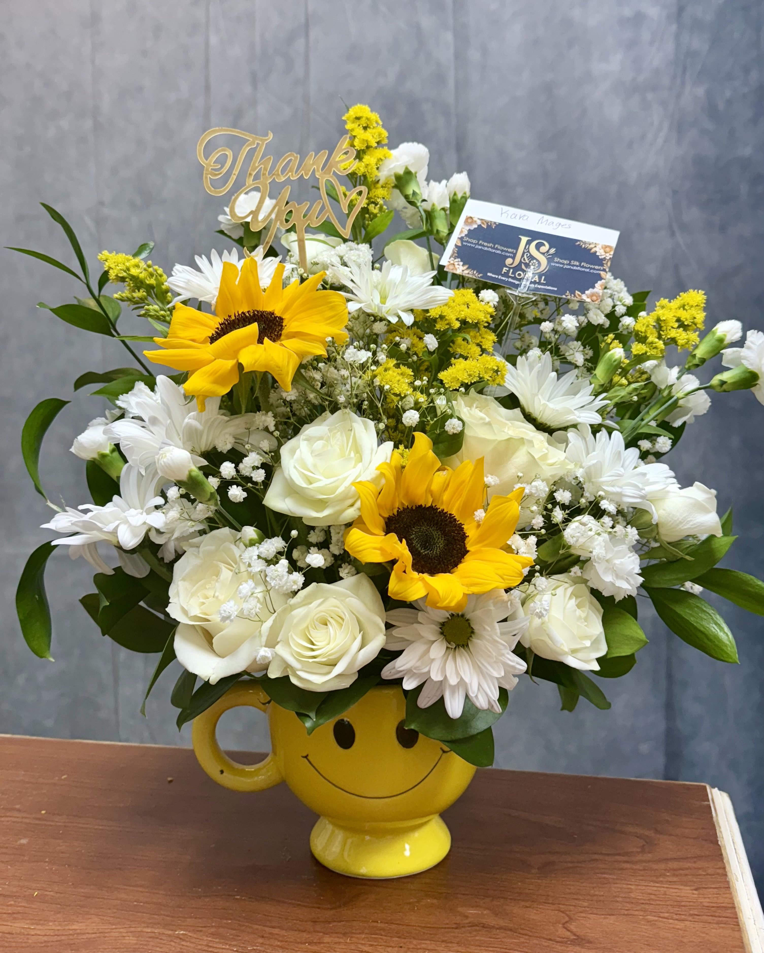 Cheerful bouquet of sunflowers, white roses, and daisies in a yellow smiley mug with a thank you pick.