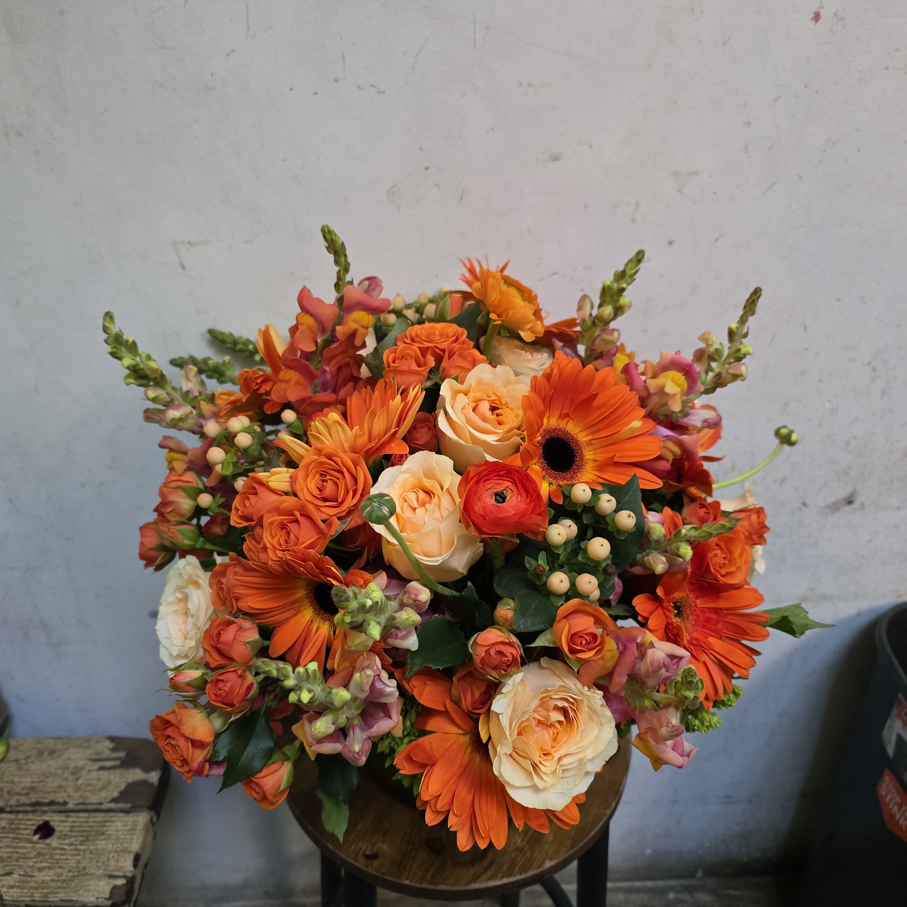 Los feliz sunset - Vivid orange gerbera daisies and coral-peach roses create an immediate sunlit impact in this Los Feliz Sunset flower arrangement-perfect as a birthday bouquet, congratulations gift, or a bright seasonal surprise. The energetic mix of warm oranges, pinks, and soft peaches reads joyful and celebratory, so the person you're thinking of will feel noticed, cheered, and truly celebrated.  Dense, rounded and garden-rich in texture, the arrangement pairs layered ranunculus-style blooms and spray roses with tall snapdragon spikes for height, plus pops of white and cream roses to soften the palette. Dark foliage and small berry-like accents add contrast while a dark vase grounds the composition for a modern, autumnal-meets-spring look. Sized to make a bold tabletop statement, it's an abundant, hand-tied style that arrives ready to display and bring instant warmth to any room.