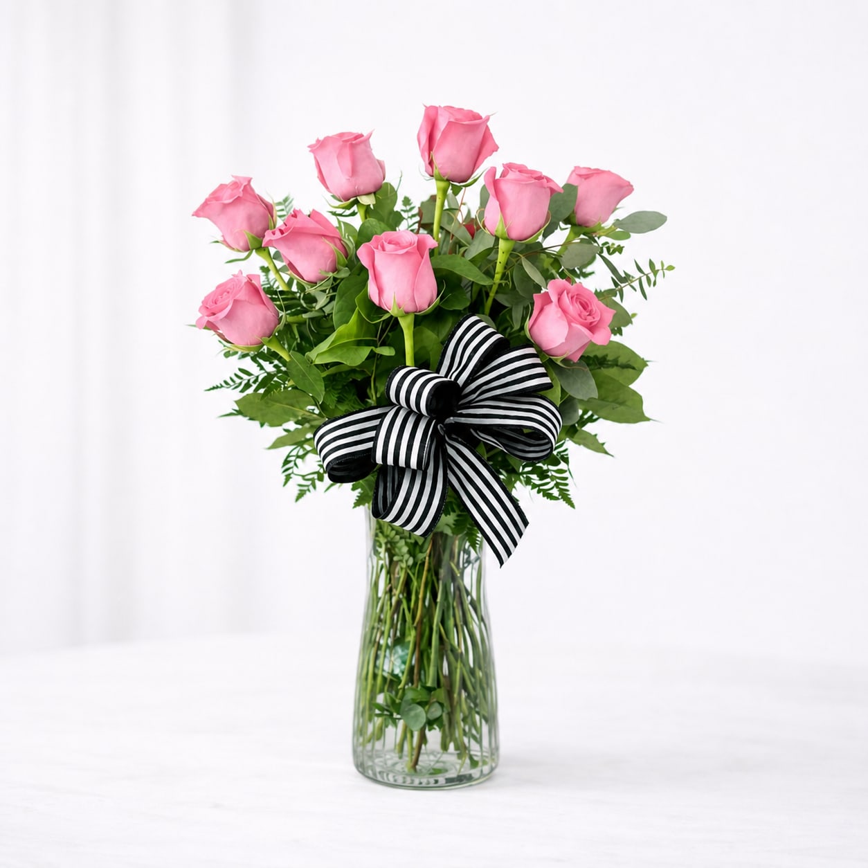 One Dozen Pink Roses - Say it beautifully with a timeless dozen roses. This elegant arrangement features twelve fresh pink roses, artfully designed with lush greenery and accented with a stylish ribbon in a clear glass vase. Perfect for birthdays, anniversaries, or just because, this classic bouquet delivers a soft, romantic feel that never goes out of style.