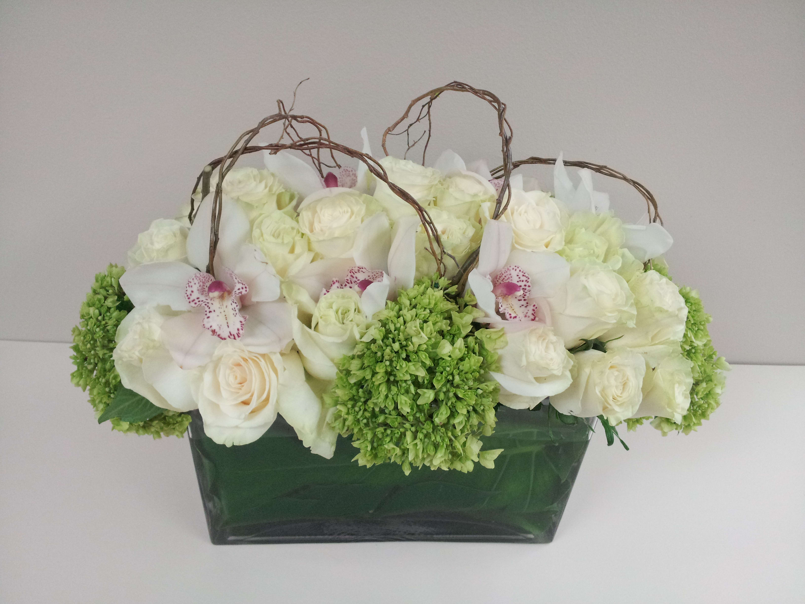 White roses and orchids arranged in a green vase with twig handles