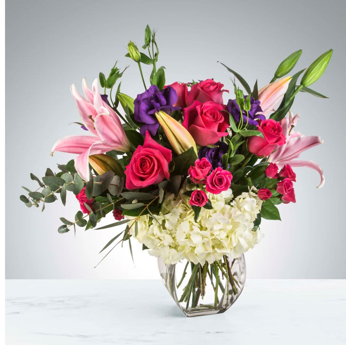 Elegance in Bloom - Transform your space with our exquisite arrangement featuring oriental pink lilies, vibrant hot pink roses, lush eucalyptus, rich purple carnations, and pristine white hydrangeas, all artfully displayed in a sophisticated vase. This luxurious bouquet not only enhances your decor but also evokes a sense of refined beauty, making it the perfect centerpiece for any occasion