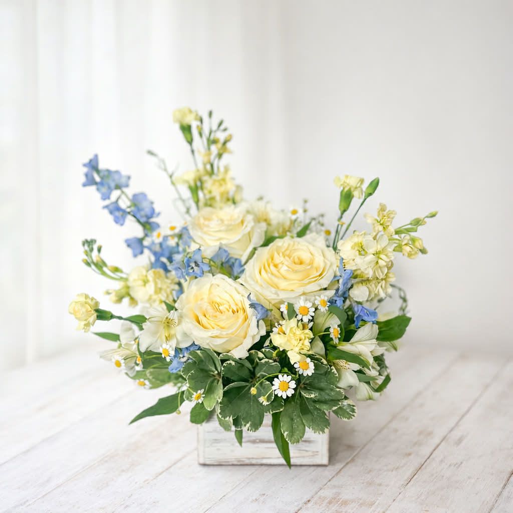 Morning Sun - Soft, fresh, and effortlessly charming—Sweet Meadow Morning captures the feeling of a peaceful spring day. Featuring buttery yellow roses, delicate blue delphinium, petite daisies, and light seasonal blooms, this arrangement is designed with a natural, garden-picked style. Arranged in a rustic wooden box, it brings a cozy, farmhouse touch that fits beautifully into any space. Perfect for sending a little sunshine, celebrating a new beginning, or simply brightening someone’s day.