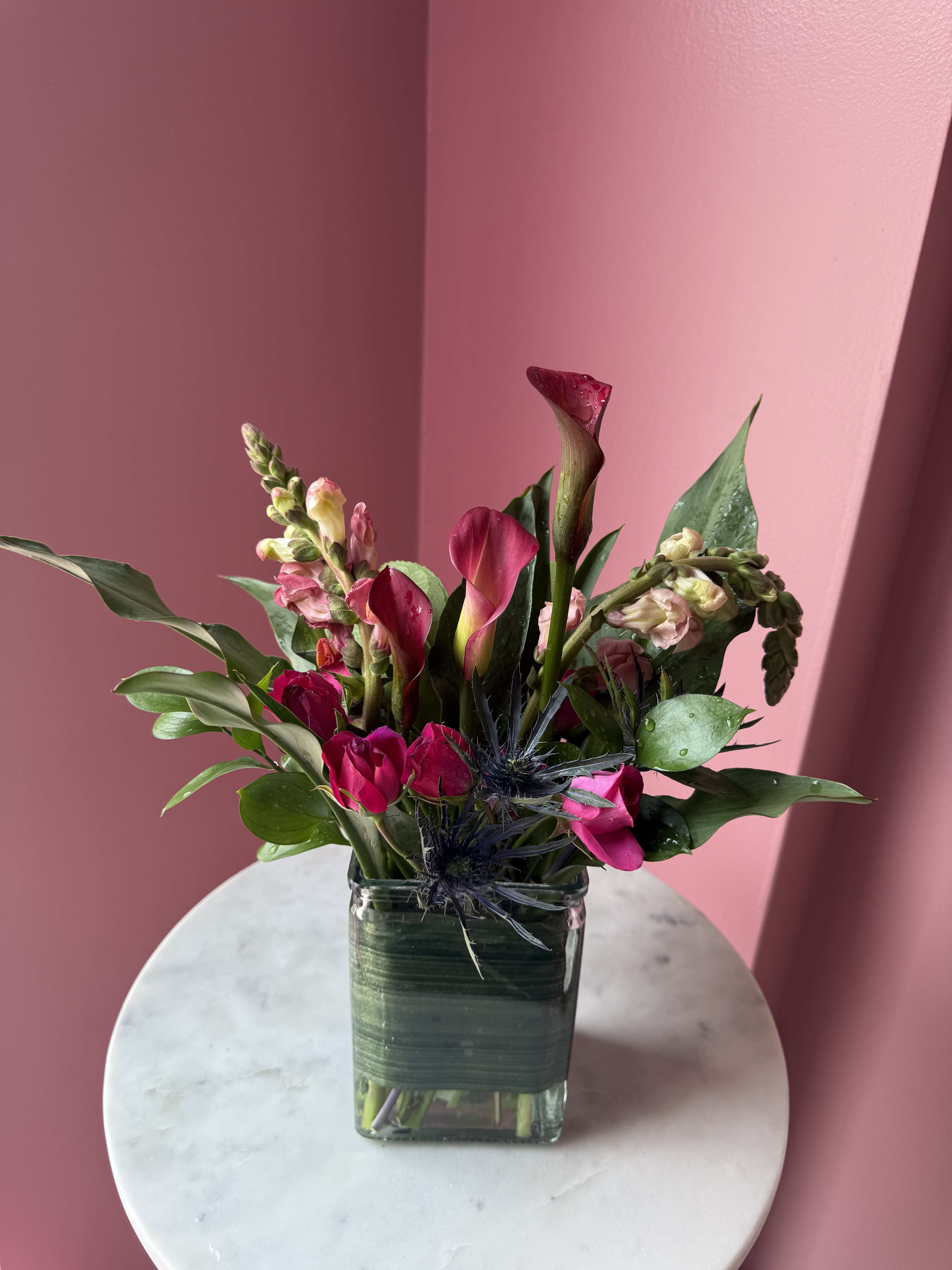 VIOLET NO. 1 - A striking, vertical composition featuring deep magenta calla lilies, purple and pink snapdragons, red alstroemeria, and a touch of textural blue thistle for contrast. Lush green foliage frames the blooms, creating a bold, architectural silhouette inside 