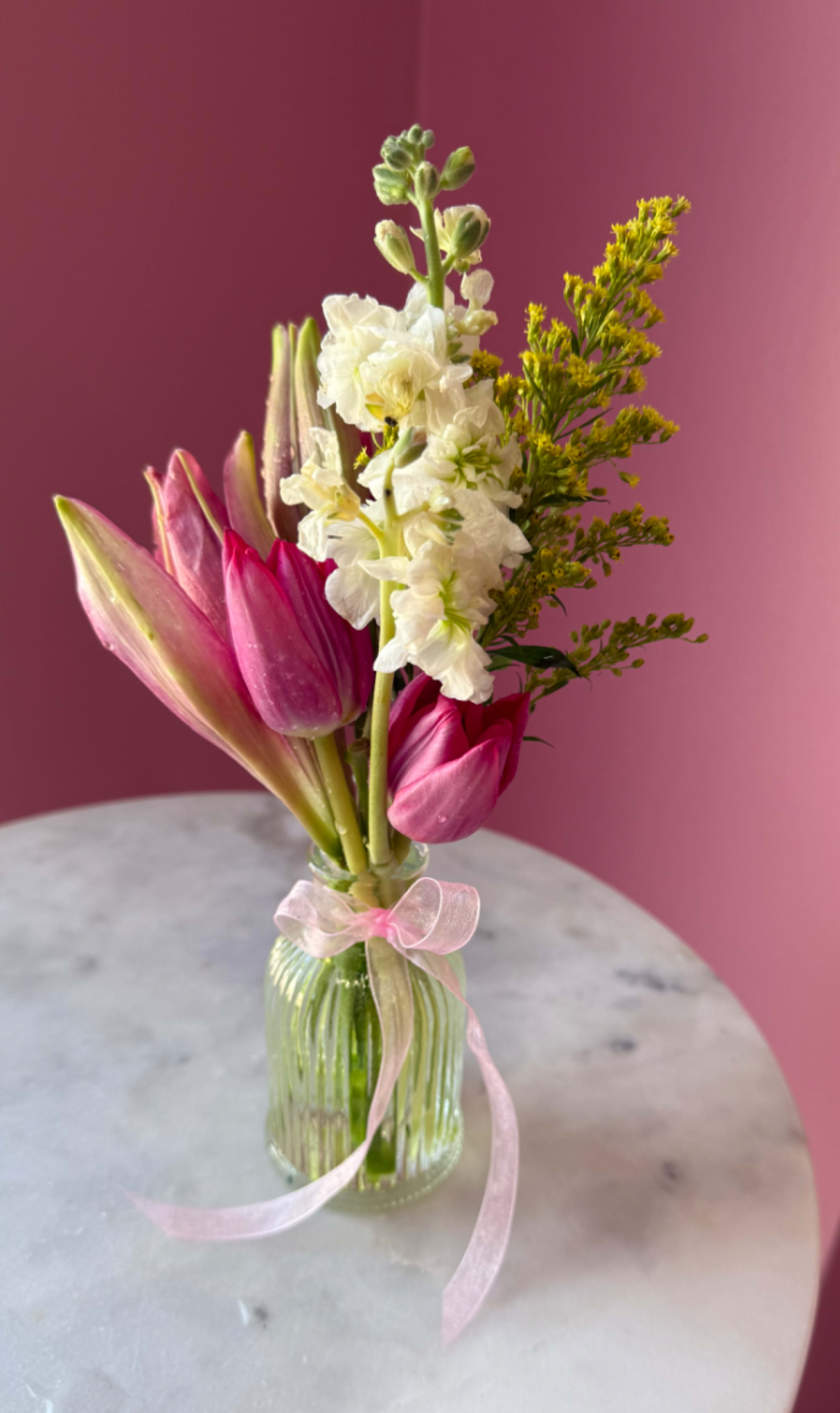 Spring Muse  - Soft pink tulips mingle with upright white stock and airy yellow-green filler to create a spring-ready bouquet that feels fresh and intimate - ideal as a Mother's Day gift, birthday surprise, thank-you arrangement, or a cheerful "just because" present. The compact, vertical silhouette makes the recipient feel genuinely seen: delicate, romantic, and quietly joyful without overwhelming a countertop or bedside table.  Gathered in a small clear ribbed glass vase and finished with a sheer pink ribbon, this arrangement balances saturated pink tulip heads and buds with pale white blooms and light green accents for a layered, textural look. The piece reads fresh and airy in person; its modest size and upright form make it perfect for casual everyday displays, small tabletops, or a thoughtful gift that brightens someone's day. Care: keep water fresh and trim stems every few days to extend bloom life.