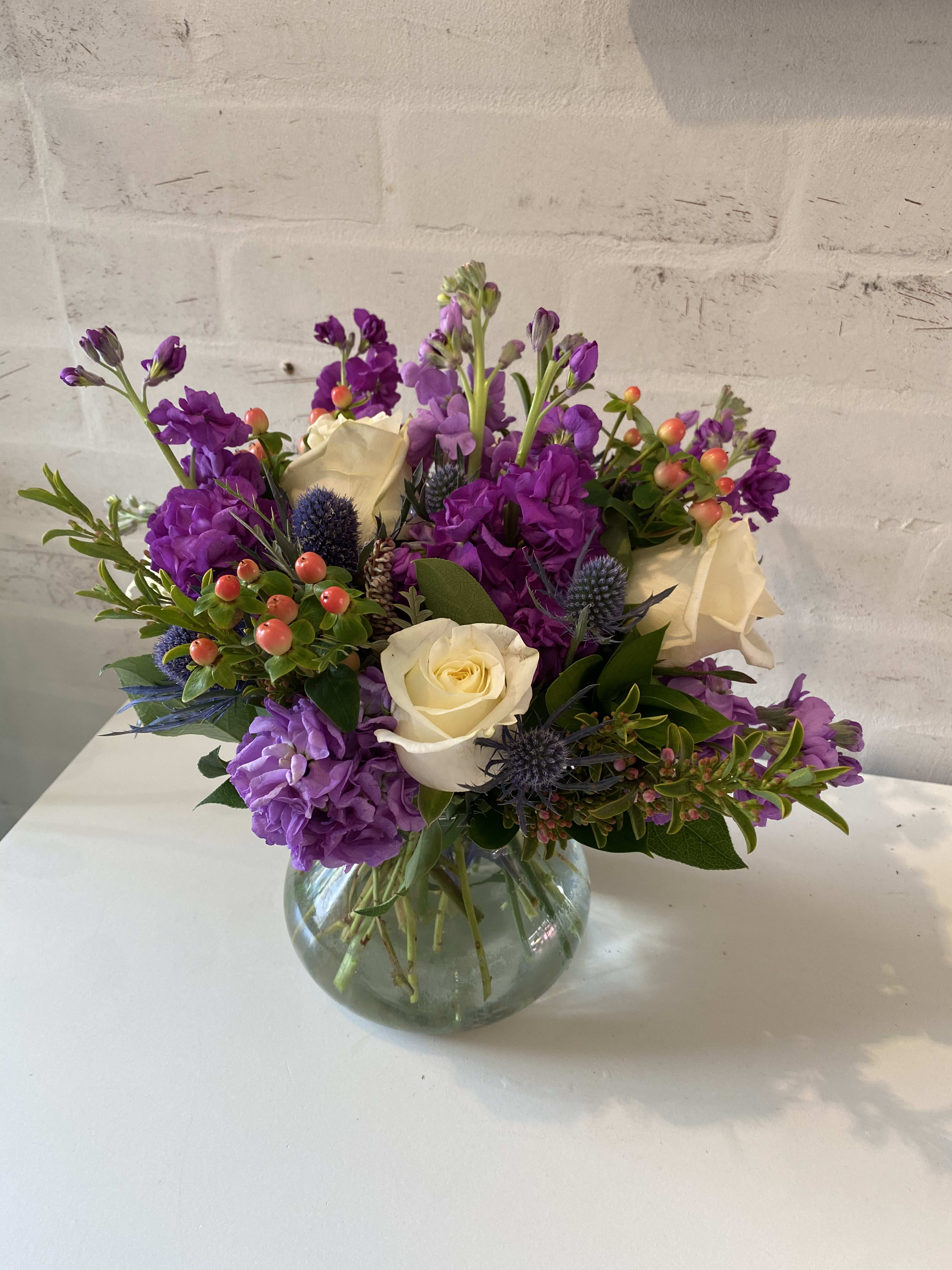 Round glass vase of white roses, purple flowers, blue thistles, and orange berries.