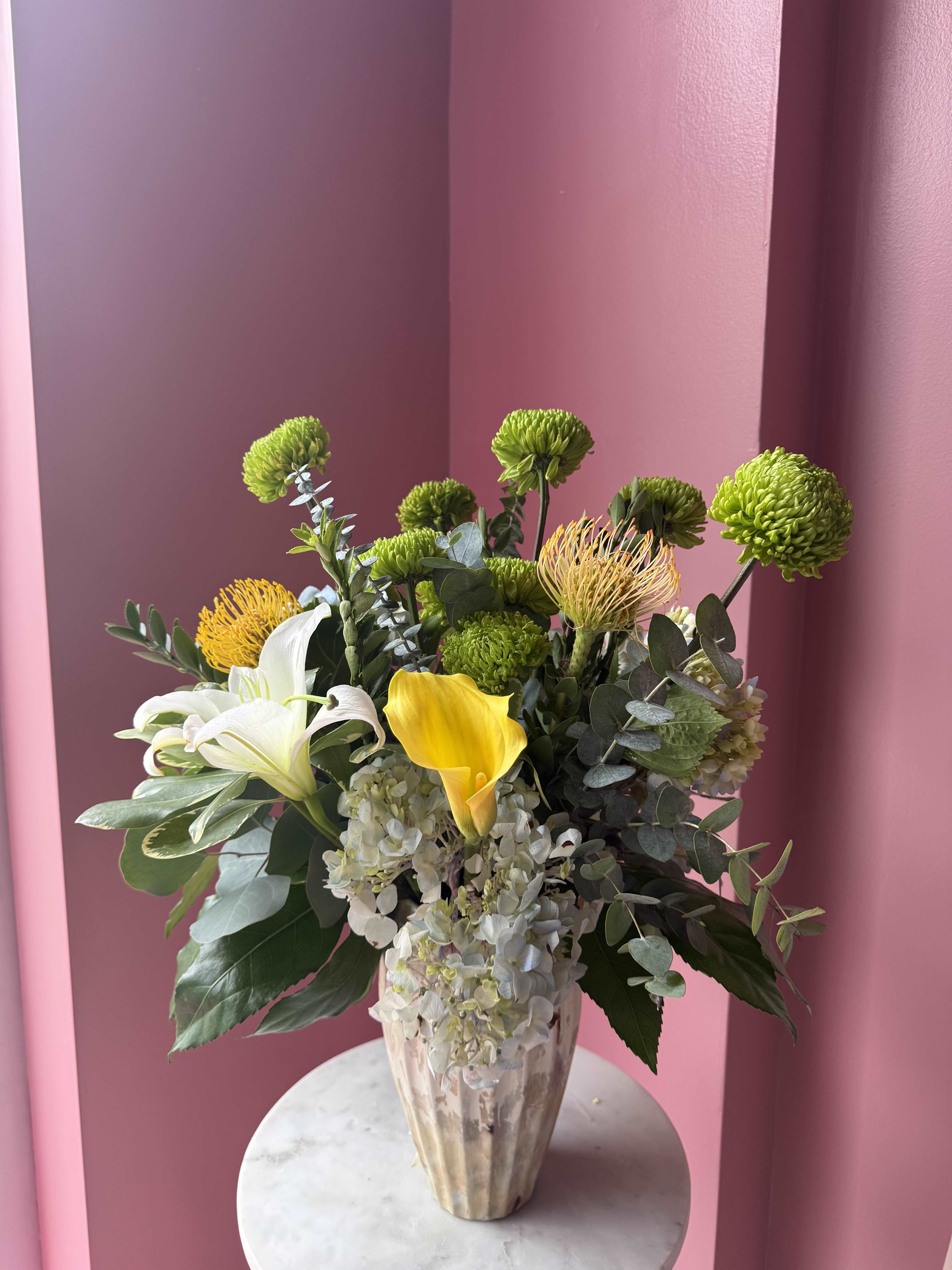 Modern Eden  - A fresh, garden-inspired centerpiece that feels both modern and effortless-the Modern Eden flower arrangement makes a lovely birthday bouquet, thank-you gift, or bright spring celebration piece. The first impression is its lively mix of chartreuse-green chrysanthemums and the sculptural height of white lilies, balanced by a single vivid yellow calla lily and soft, pale hydrangea clusters so the person you're thinking of will feel genuinely celebrated and seen.  This full, layered arrangement pairs rounded blooms and broad foliage with a spiky protea-like accent for contrast and texture. Colors include green, white, yellow, and touches of pink from the background tones; flowers featured are lilies, calla lilies, chrysanthemums, hydrangeas, and protea-style blooms, all presented in a clear glass vase that highlights the clean, contemporary silhouette.  Perfect for brightening a table, office, or entryway, Modern Eden reads as an everyday yet memorable gift that says "you're on my mind" without being formal. The design arrives ready to display in the glass vase-trim and change the water every few days to keep the stems fresh-and choose a size that fits the space you have in mind.