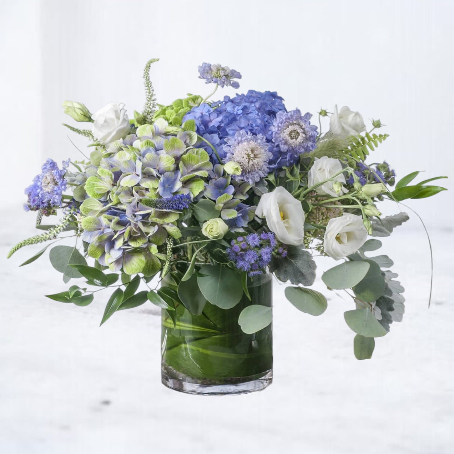 Cloudbank Drift - Cloudbank Drift is an airy, garden-style floral arrangement designed with soft movement, layered texture, and a refined cool-toned palette. Featuring premium blue hydrangea, soft green hydrangea, white lisianthus, and delicate lavender accent blooms, this design is artfully arranged with fresh eucalyptus and seasonal greenery for a naturally flowing composition.  Designed in a modern glass vessel with lush greenery wrapping the base, Cloudbank Drift blends cool blues, soft greens, and crisp whites to create a serene, elevated look. The combination of hydrangea and airy accent florals gives this arrangement a light, cloud-like presence while maintaining a full, luxurious feel.  Perfect for sympathy flowers, get well wishes, birthdays, or sending a calming, elegant gift, this blue and green flower arrangement is a standout choice for those drawn to soft, nature-inspired designs.  Same-day flower delivery is available in Magnolia, TX, The Woodlands, Montgomery, Conroe, Spring, Tomball, and surrounding North Houston areas.  Due to seasonal availability, some blooms may be replaced with similar varieties of equal or greater value to maintain the design’s look and integrity. Your arrangement will always reflect the same color palette, style, and premium quality.