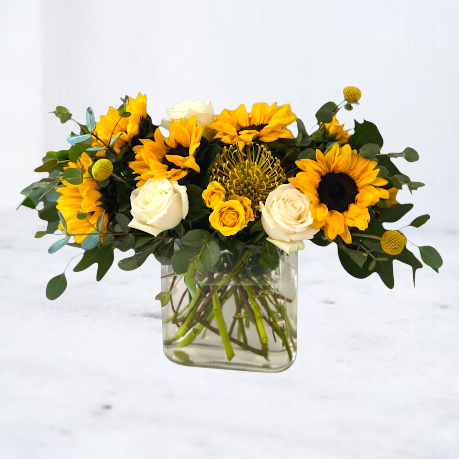 Solaris Charm - Solaris Charm is a radiant, design-forward floral arrangement that captures the warmth of golden hour in bloom. Featuring vibrant sunflowers, creamy white roses, golden billy balls, and textured pincushion protea, this composition is layered with fresh eucalyptus and seasonal greenery for a modern, airy finish. Designed in a modern glass vase, Solaris Charm blends bold color with soft neutrals, creating a balanced look that feels both elevated and effortlessly natural. The contrast of rich yellow tones against crisp whites makes this arrangement perfect for brightening homes, celebrating milestones, or sending a thoughtful, uplifting gift.  Ideal for birthdays, congratulations, thank-you gestures, or just because, this sunflower and rose arrangement delivers a fresh, contemporary aesthetic that stands out from traditional floral designs.  Same-day flower delivery is available in Magnolia, TX, The Woodlands, Montgomery, Conroe, Spring, Tomball, and surrounding North Houston areas.