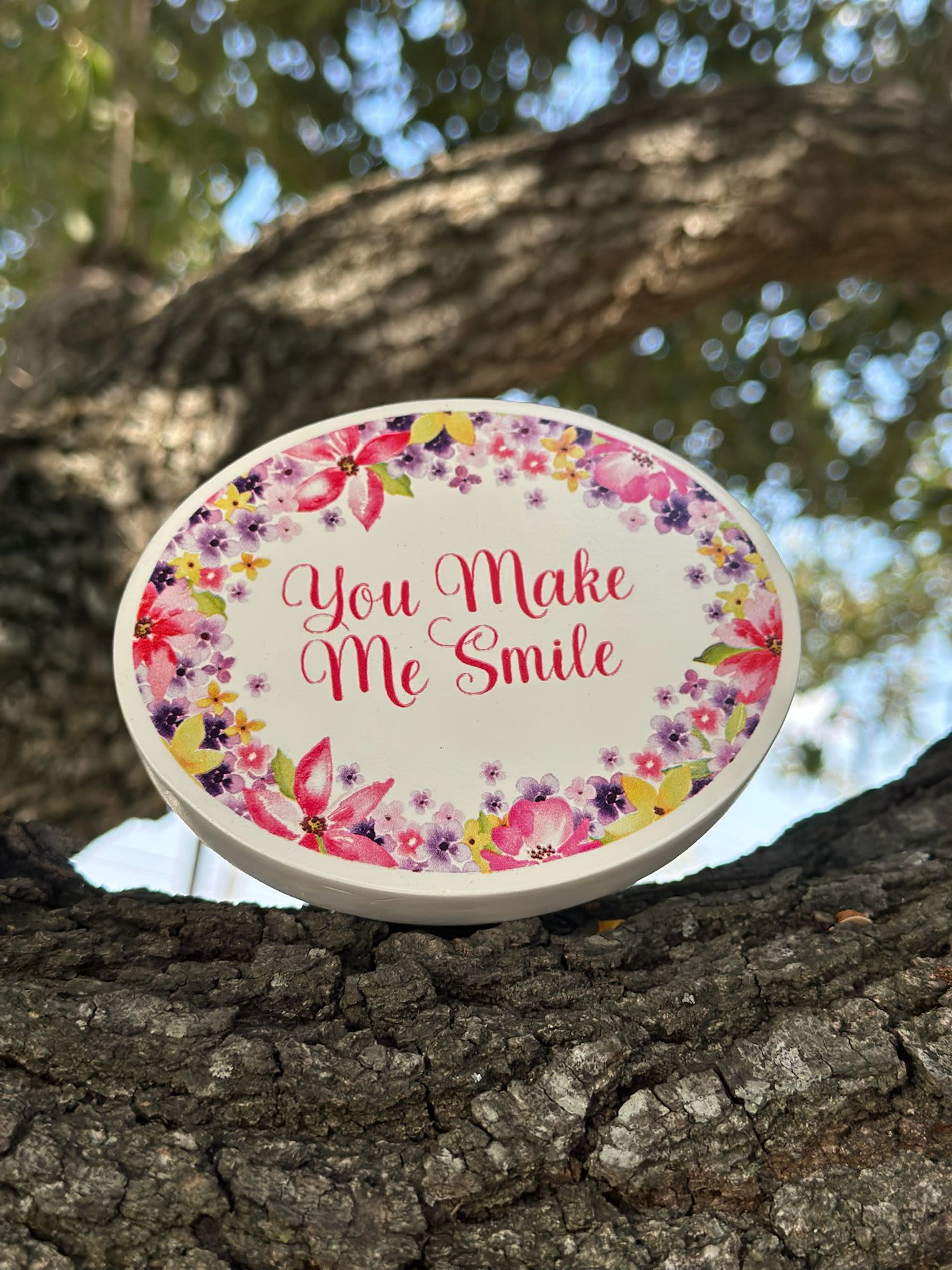 Oval floral sign reading "You Make Me Smile" on a tree branch