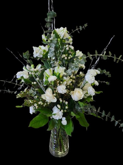 Airy Medow - Tall white roses and alstromelia, hypericum, wax flowers create an elegant centerpiece for a sympathy arrangement, thank-you gift, or graceful housewarming bouquet. The open, airy silhouette-lifted by eucalyptus branches that extend beyond the bloom line-gives the recipient a sense of calm and thoughtful presence, the kind of arrangement that quietly says you care.  This flower arrangement pairs pure white roses, alstromelia,  and carnations with small white filler blooms and subtle berry-like accents for texture, all arranged in a clear glass vase. The fresh green-and-white palette and varied heights make it feel light and refined; proportions read tall and open rather than dense, perfect for a mantel, entry table, or bedside. Care: keep water fresh, recut stems occasionally, and place away from direct heat to prolong blooms.