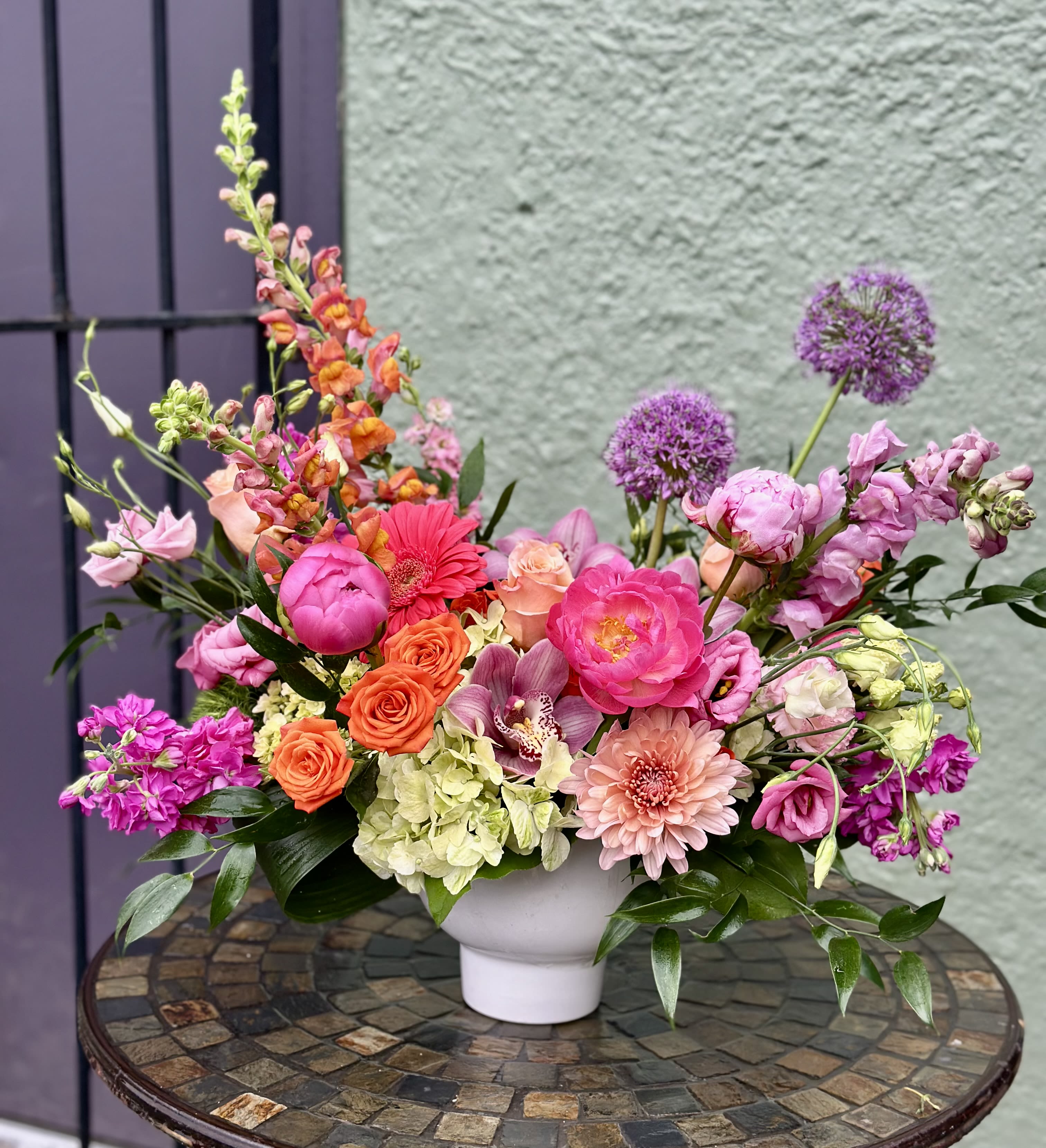 Spring Charm - A lush, garden-style centerpiece that feels like a sunlit spring morning-this flower arrangement is a cheerful bouquet in a white ceramic vase perfect for this Mother's Day gift. Large pink and peach roses and full ranunculus anchor the design, while pops of yellow spray roses and purple accents lend vibrant contrast; the overall effect is bright, feminine, and celebratory so the person you're thinking of will feel truly seen.