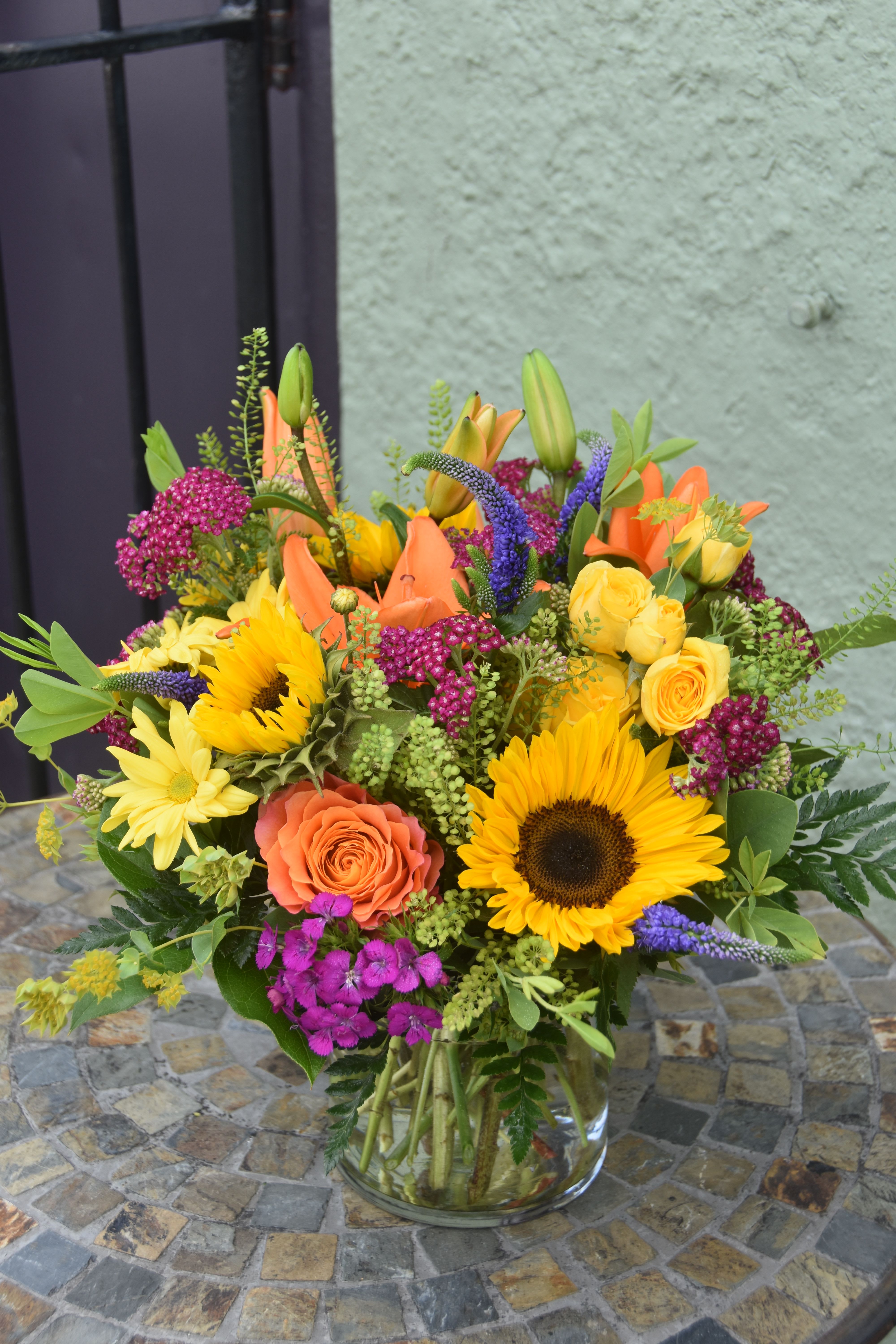 Bright mixed arrangement of sunflowers, roses, lilies and daisies in a clear glass vase