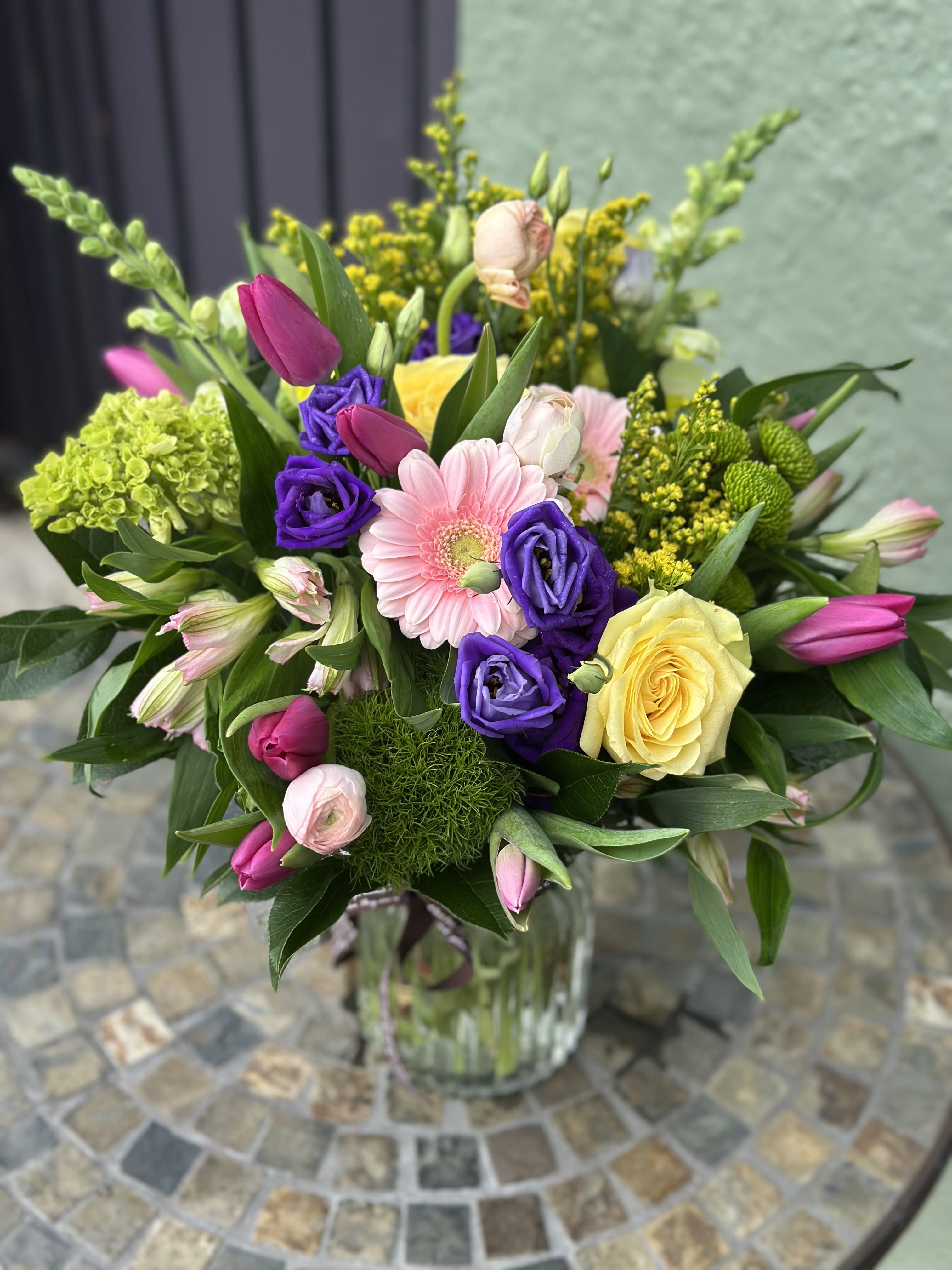 Vase arrangement of pink tulips, purple lisianthus, a yellow rose and mixed blooms in a clear glass vase.