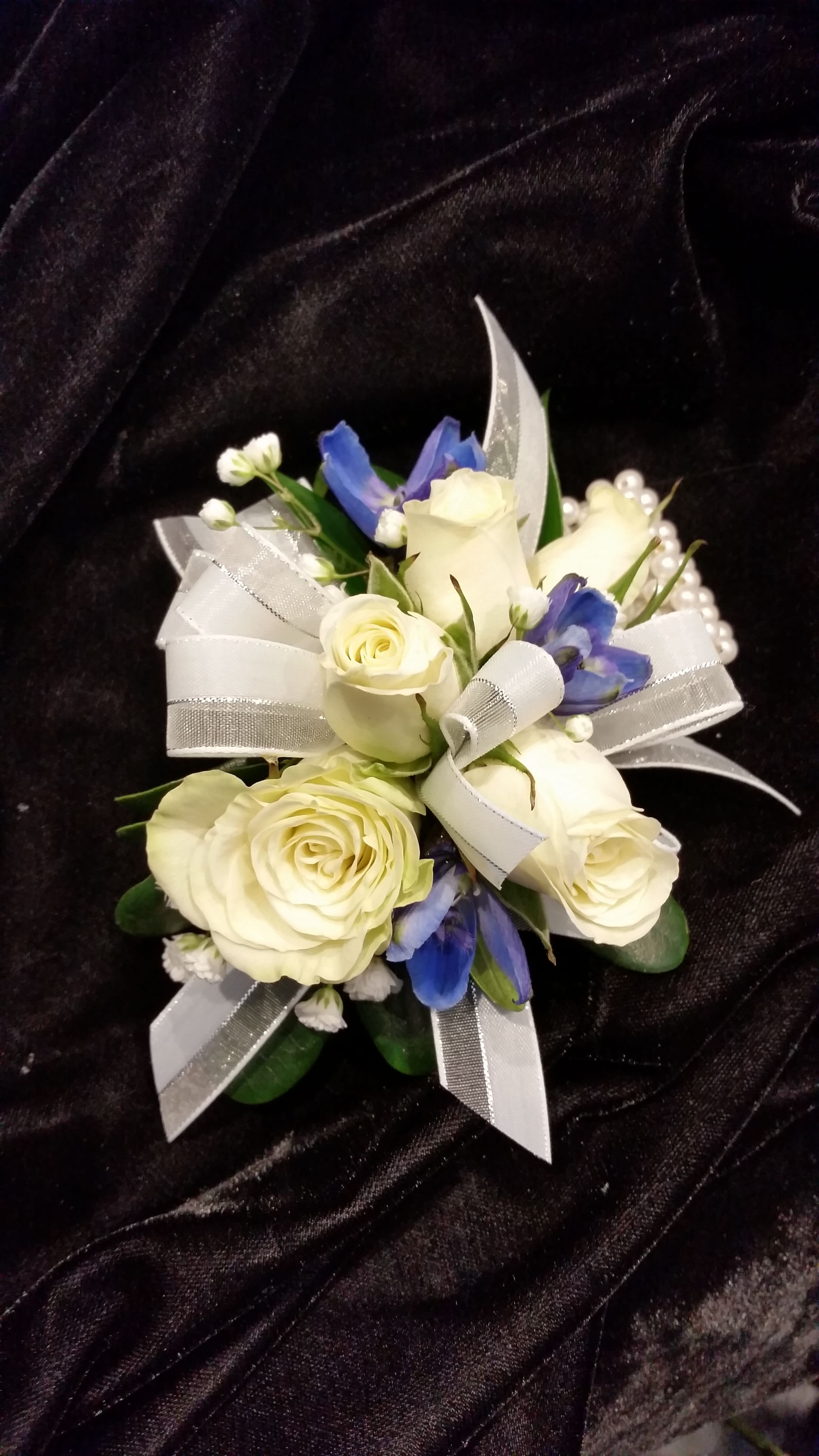Royal Wrist Corsage - One of our favorite wrist corsages features royal blue and white blooms on a pearl bracelet (pearl bracelet no longer available, we will use a standard wristlet).  Pure white spray roses or dendrobium orchid blossoms and deep blue delphinium accents create a dramatic design that works with many of the dress colors popular this year.  You can not go wrong with the Royal wrist corsage   PLEASE CALL THE SHOP AT (617)244-4422 FOR ALL CORSAGE OR BOUTONNIERE ORDERS. THANK YOU.