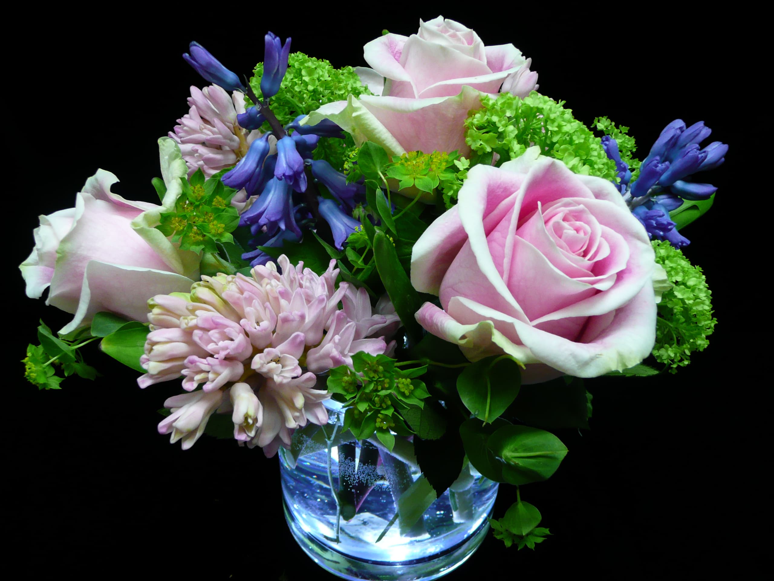 Short arrangement of pink roses, hyacinths, and green blooms in a clear glass vase on a black background