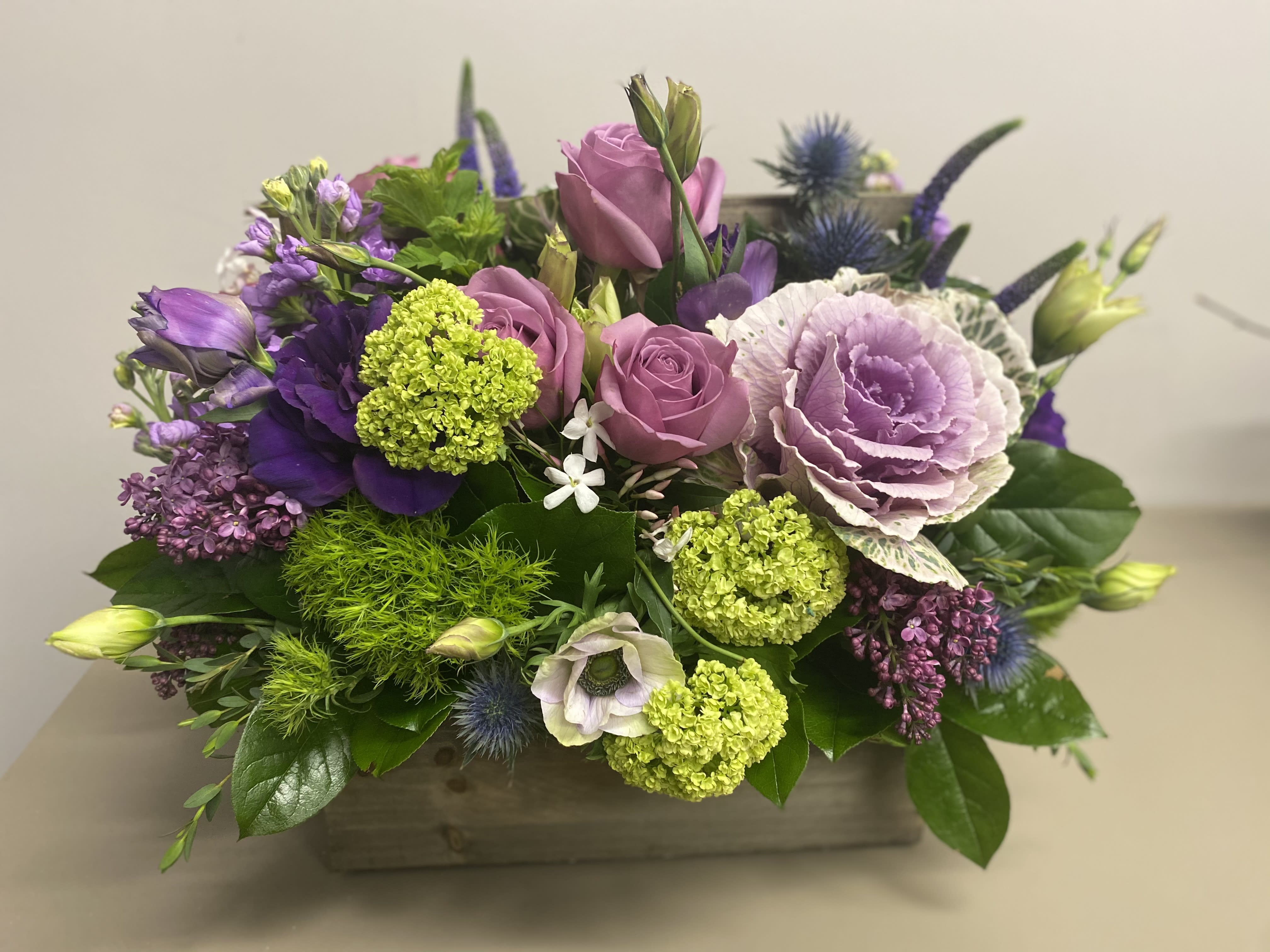 Low centerpiece of lavender roses, ornamental cabbage, green blooms and purple flowers in a wooden box