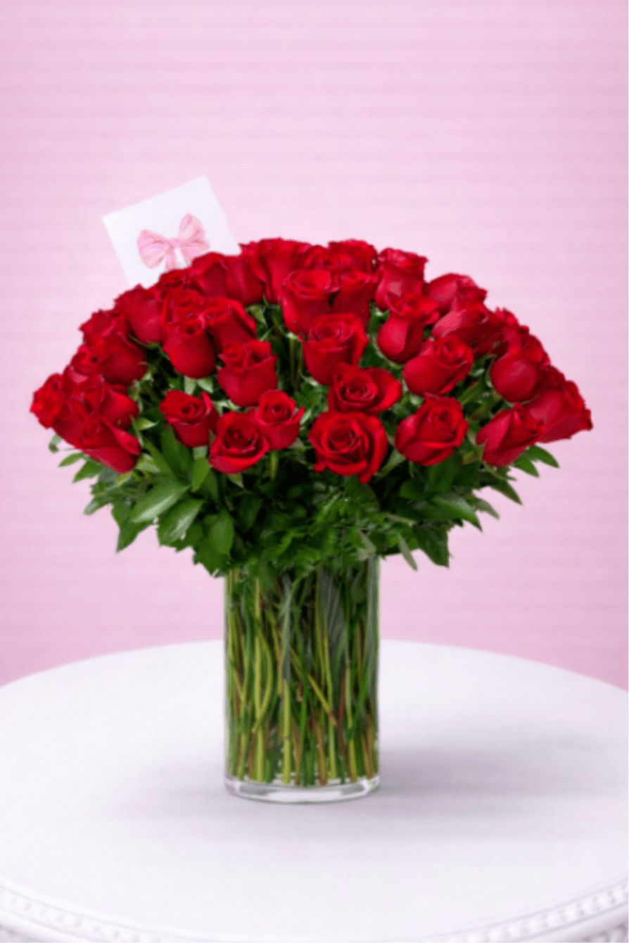 Tall clear vase filled with many red roses and a small card on a white table