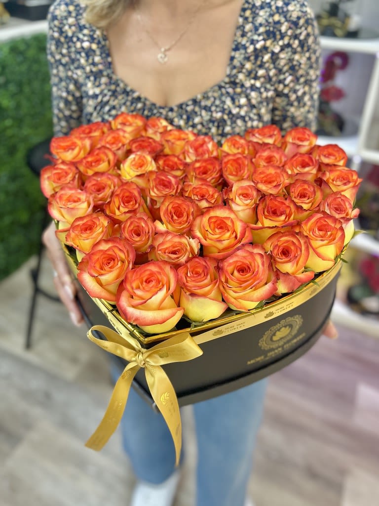 Heart-shaped black box filled with orange and yellow roses and tied with a gold ribbon
