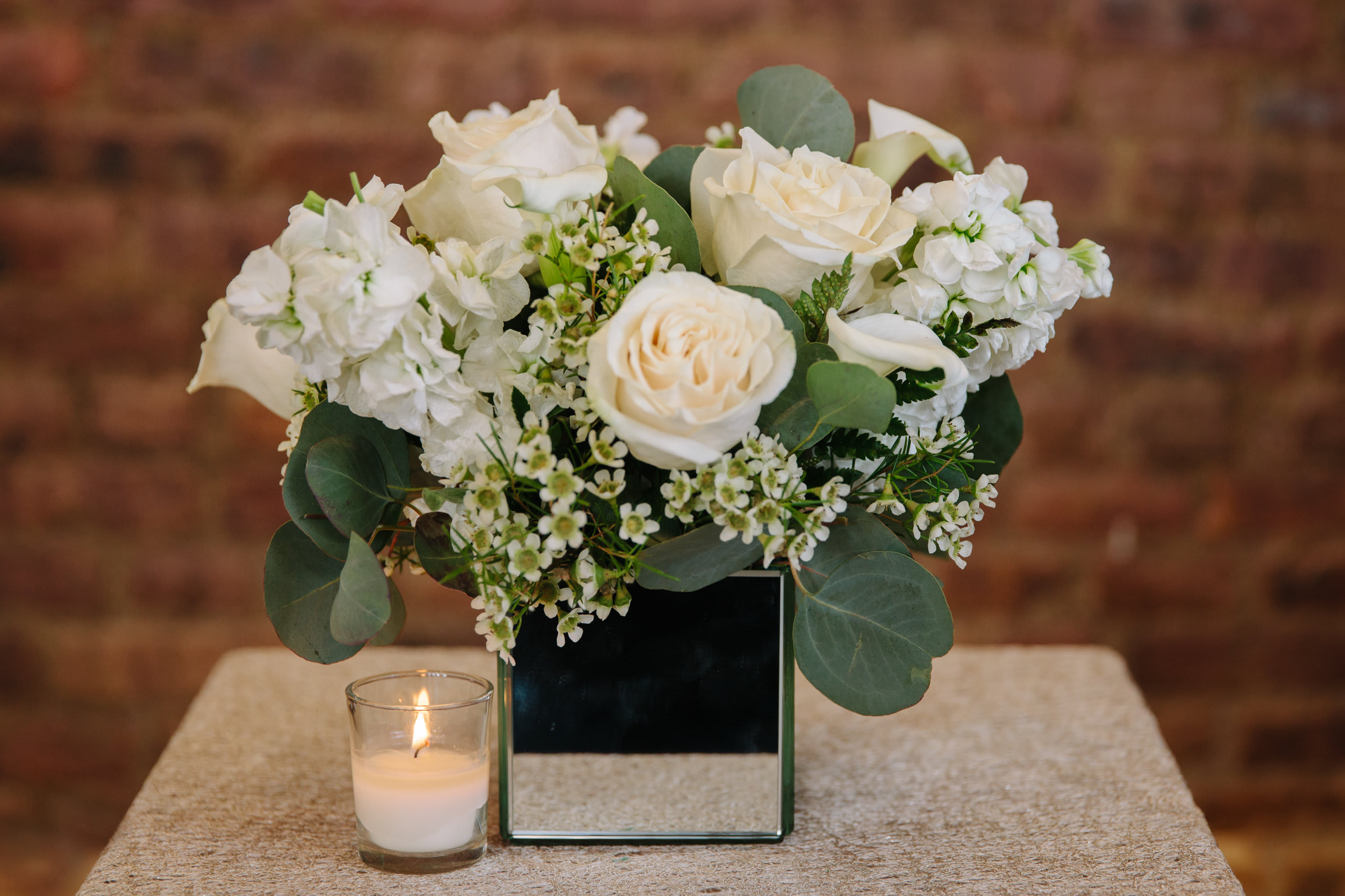 White rose arrangement in a mirrored square vase with a small candle beside it