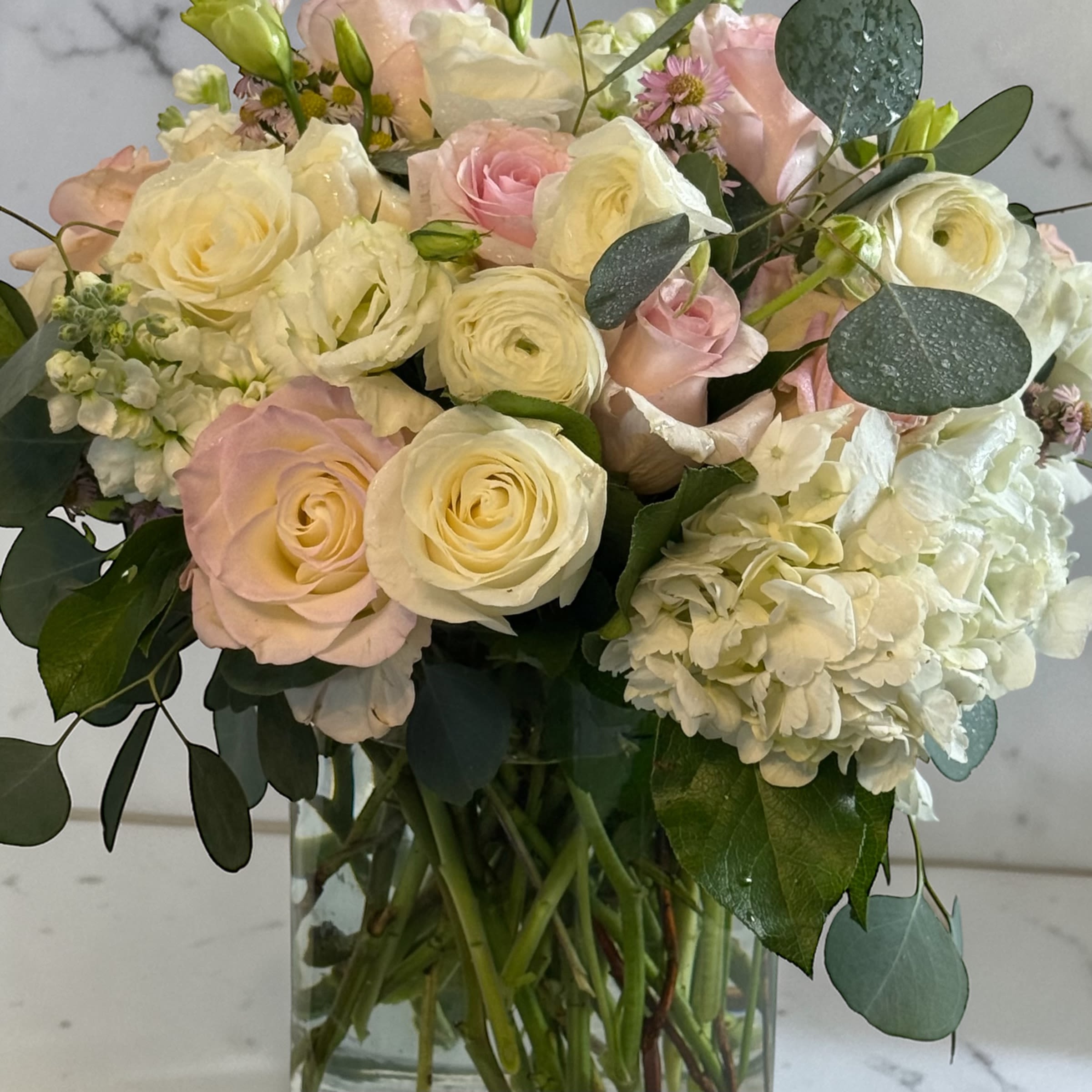 Pastel rose garden by Petal Perfection - Cream and blush roses mingle with layered white ranunculus and ruffled lisianthus in a soft pastel wedding- or anniversary-ready bouquet - a Mom's pastel rose garden designed to make the person you're thinking of feel truly cherished. Voluminous white hydrangeas and peach-toned stock add romantic texture while small asters and buds give delicate, sentimental detail; eucalyptus rounds out the palette with cool green contrast that keeps the arrangement fresh and modern.  Arranged tightly in a clear glass vase that shows the gathered stems, this compact, rounded design reads elegant on a dining table, bedside, or mantel. The mix of cream, pink, peach, and green creates a gentle, uplifting scent and a layered visual impression that looks full from every angle. Ideal for Mother's Day, birthdays, anniversaries, or as a heartfelt sympathy gesture - a thoughtful way to say "you're loved" with flowers that arrive ready to display.