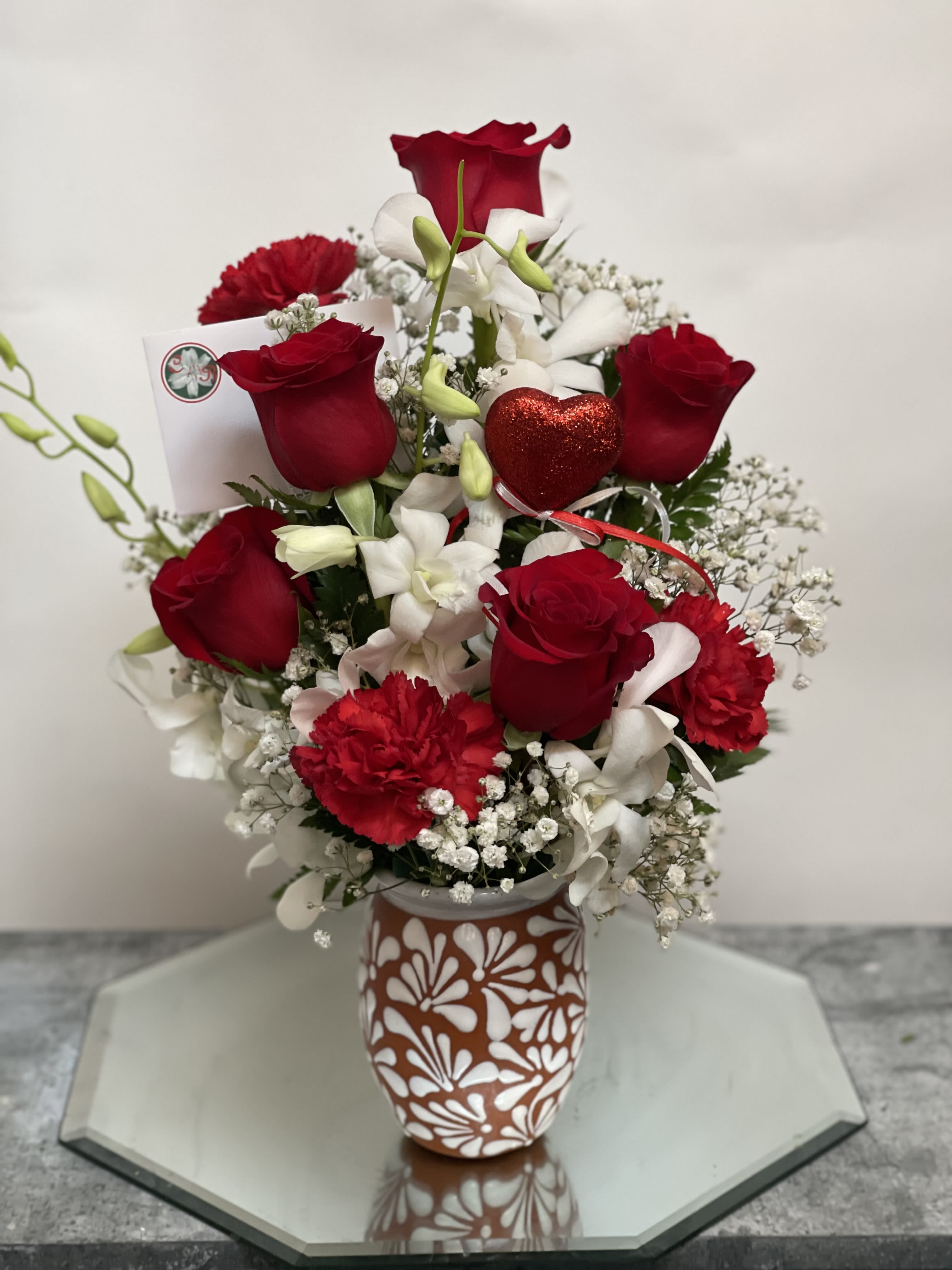 Red roses and white flowers arranged in a patterned vase with a red heart pick