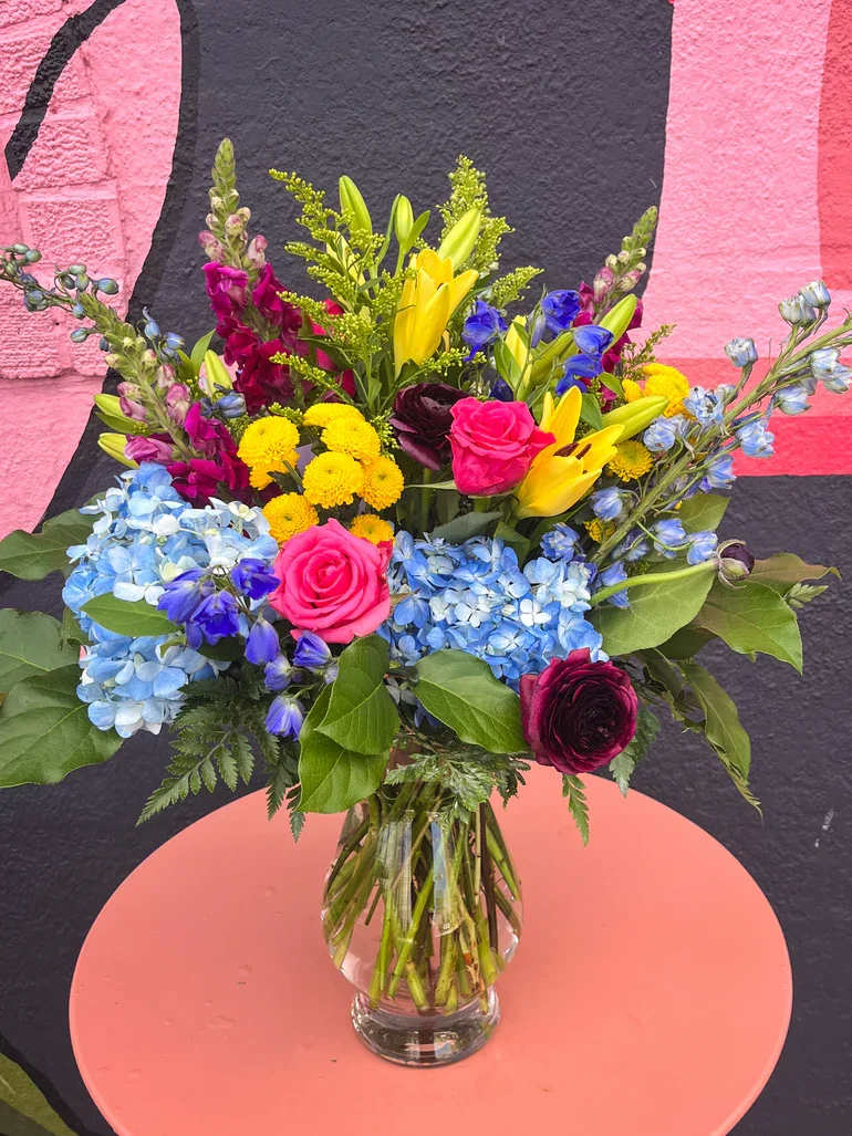 Colorful mixed bouquet with blue hydrangeas, yellow lilies, and pink roses in a clear glass vase.