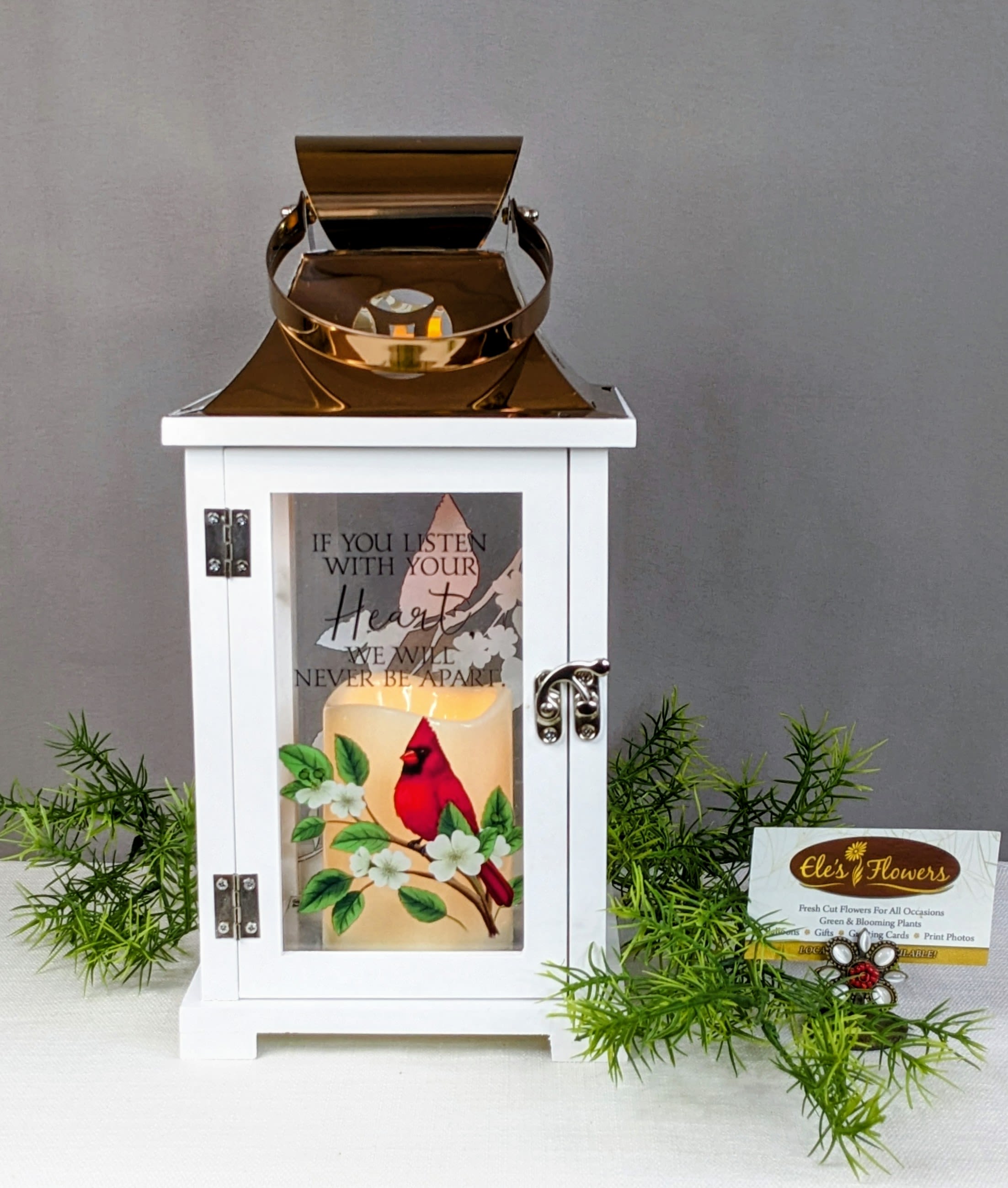 White lantern with a lit candle and cardinal design, surrounded by greenery.