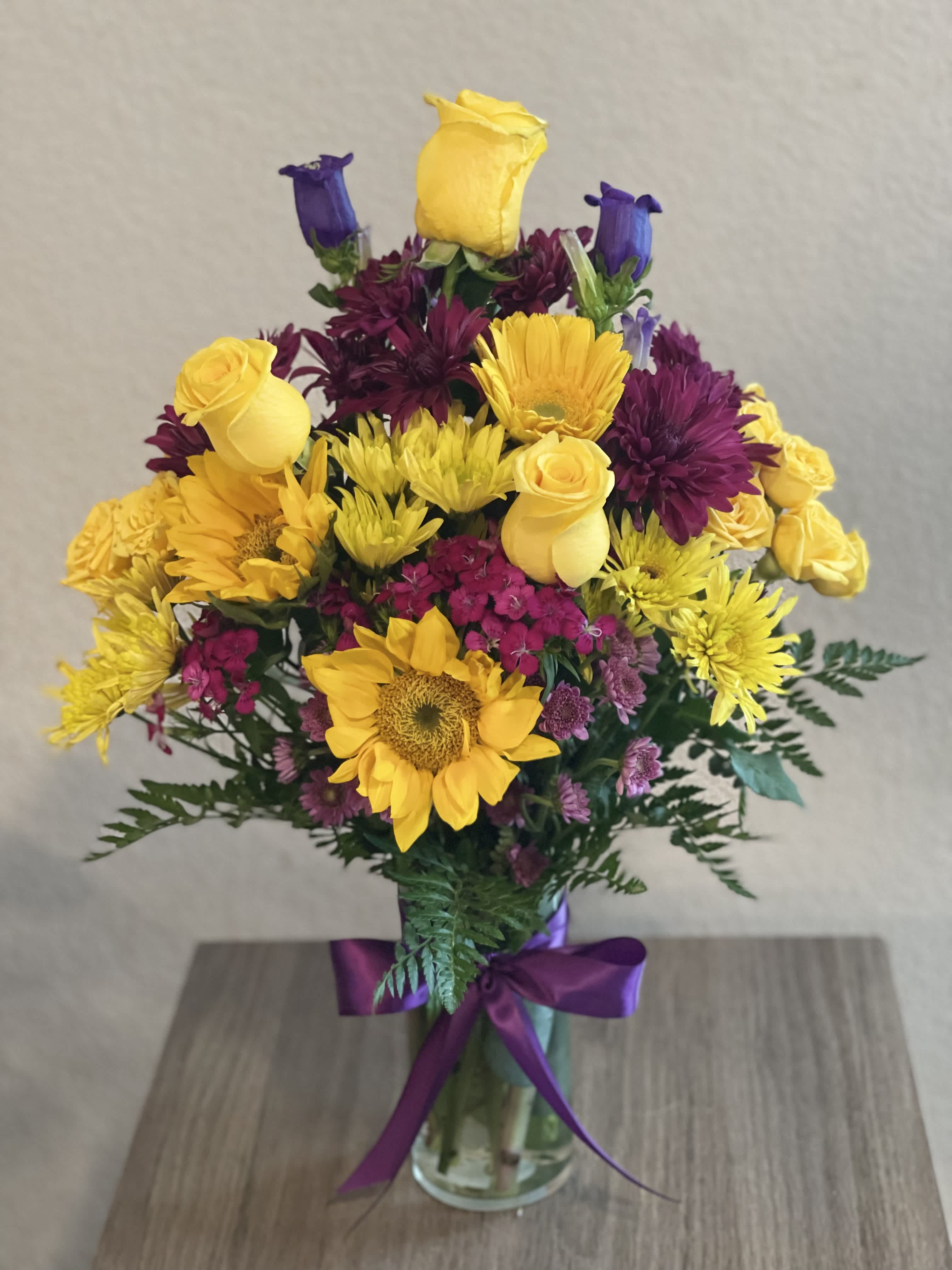 Bouquet of yellow roses, sunflowers, and purple blooms in a glass vase
