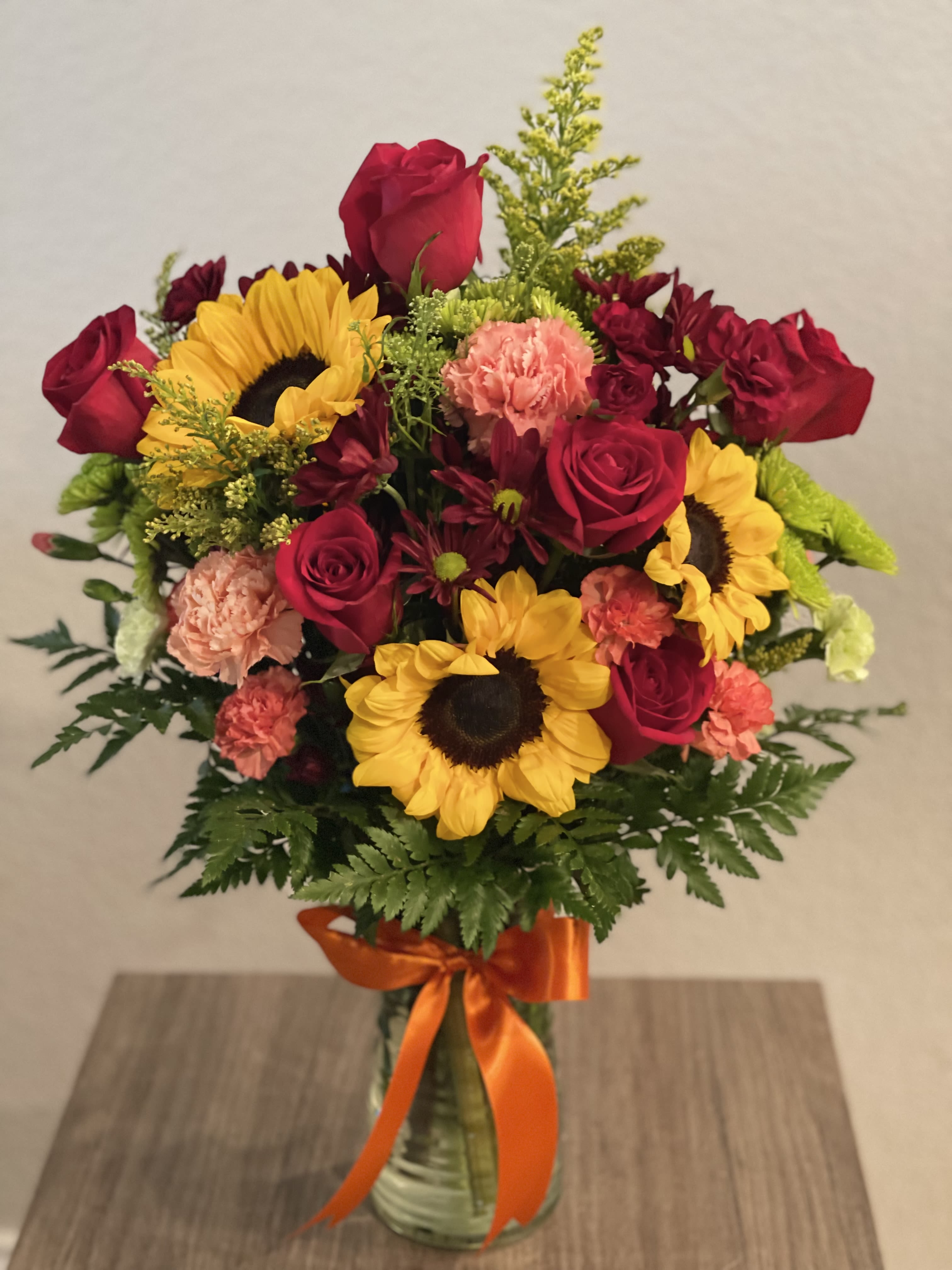 Bouquet of red roses, sunflowers, and carnations in a glass vase with orange ribbon