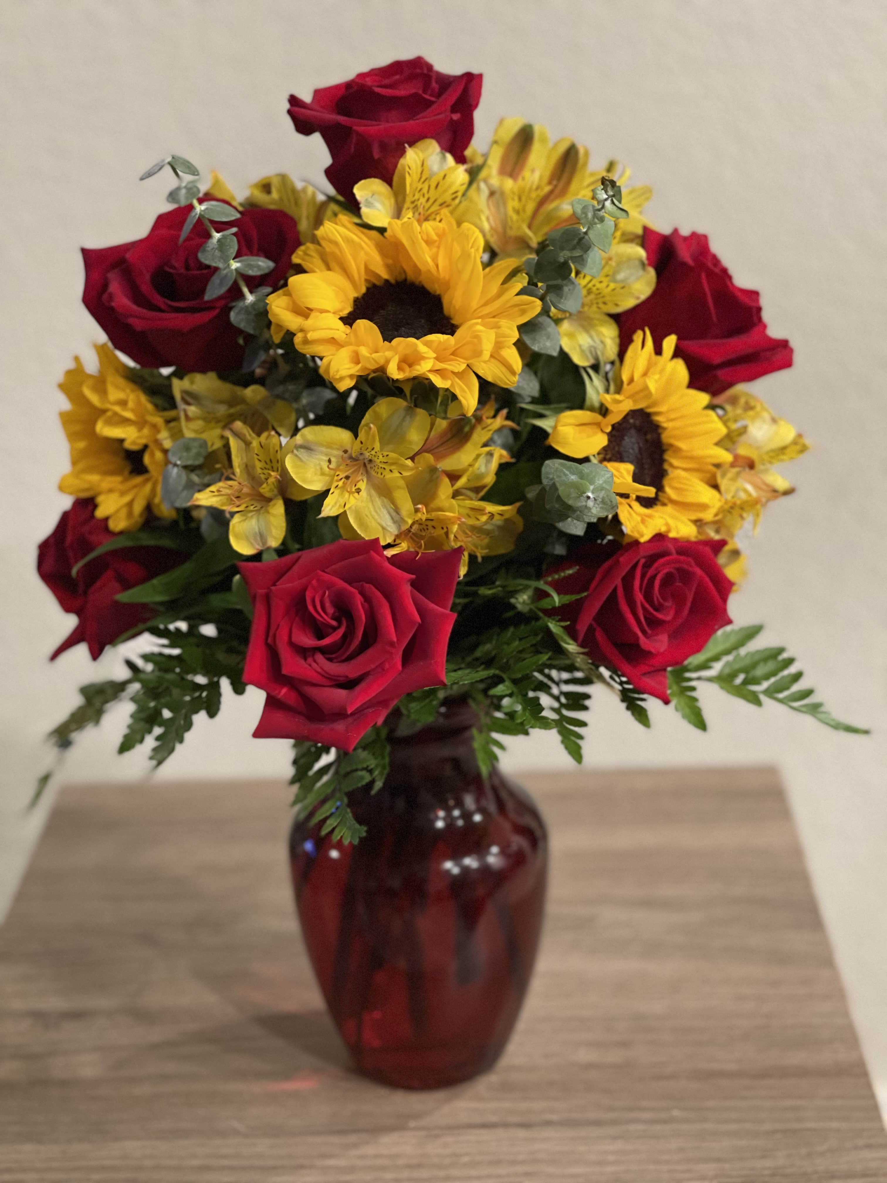Sunrise Flowers Arrangement - Crafted with care and attention to detail, this bouquet features a harmonious combination of warm red roses and delicate yellow alstroemeria flowers and sunflowers This arrangement captures the joyfulness of vibrant hues. Assorted fresh greenery adds some contrast. All the fresh blooms and greens are displayed in a glass red vase.