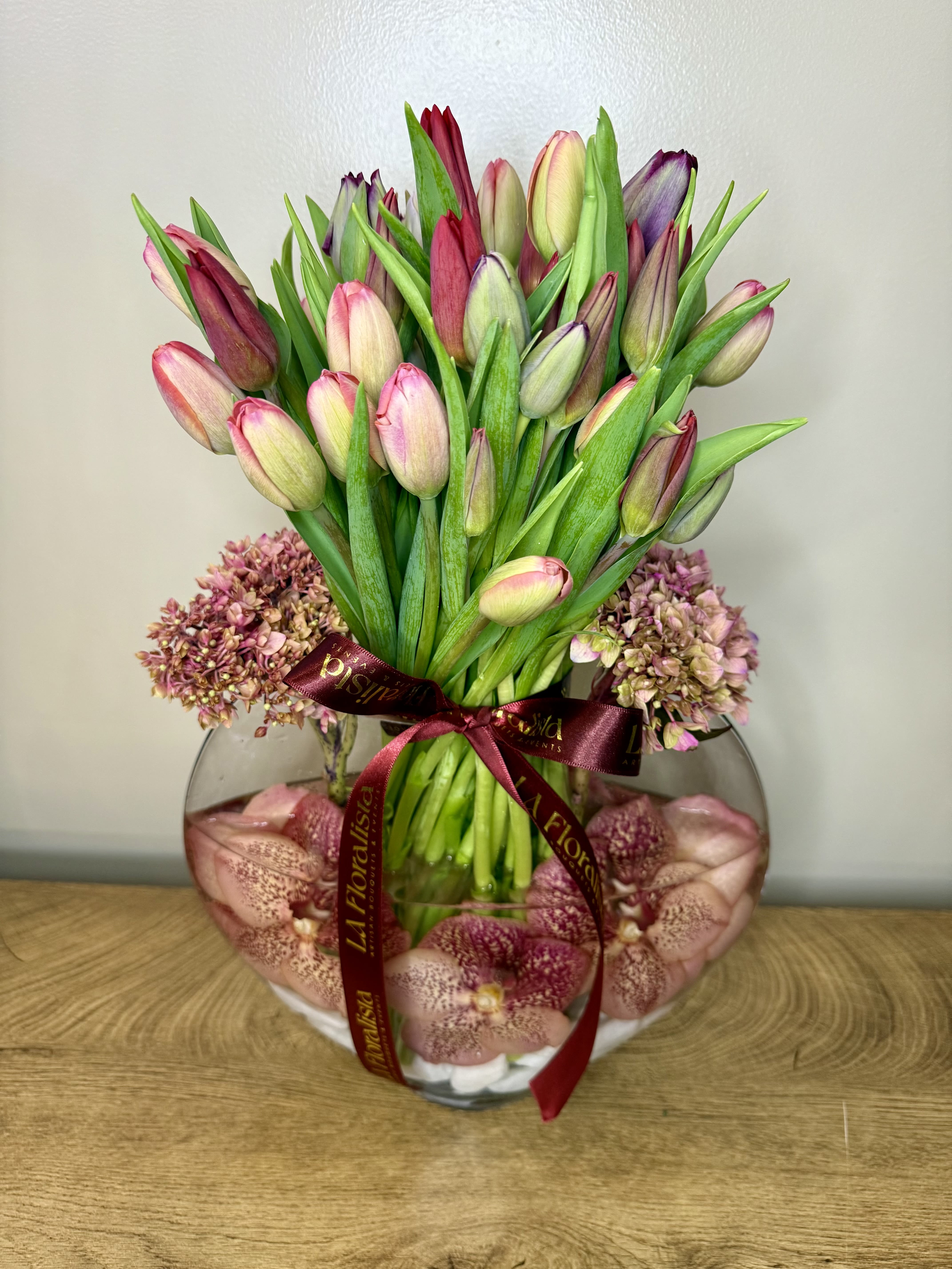 Pink and purple tulips in a round glass vase with a burgundy ribbon