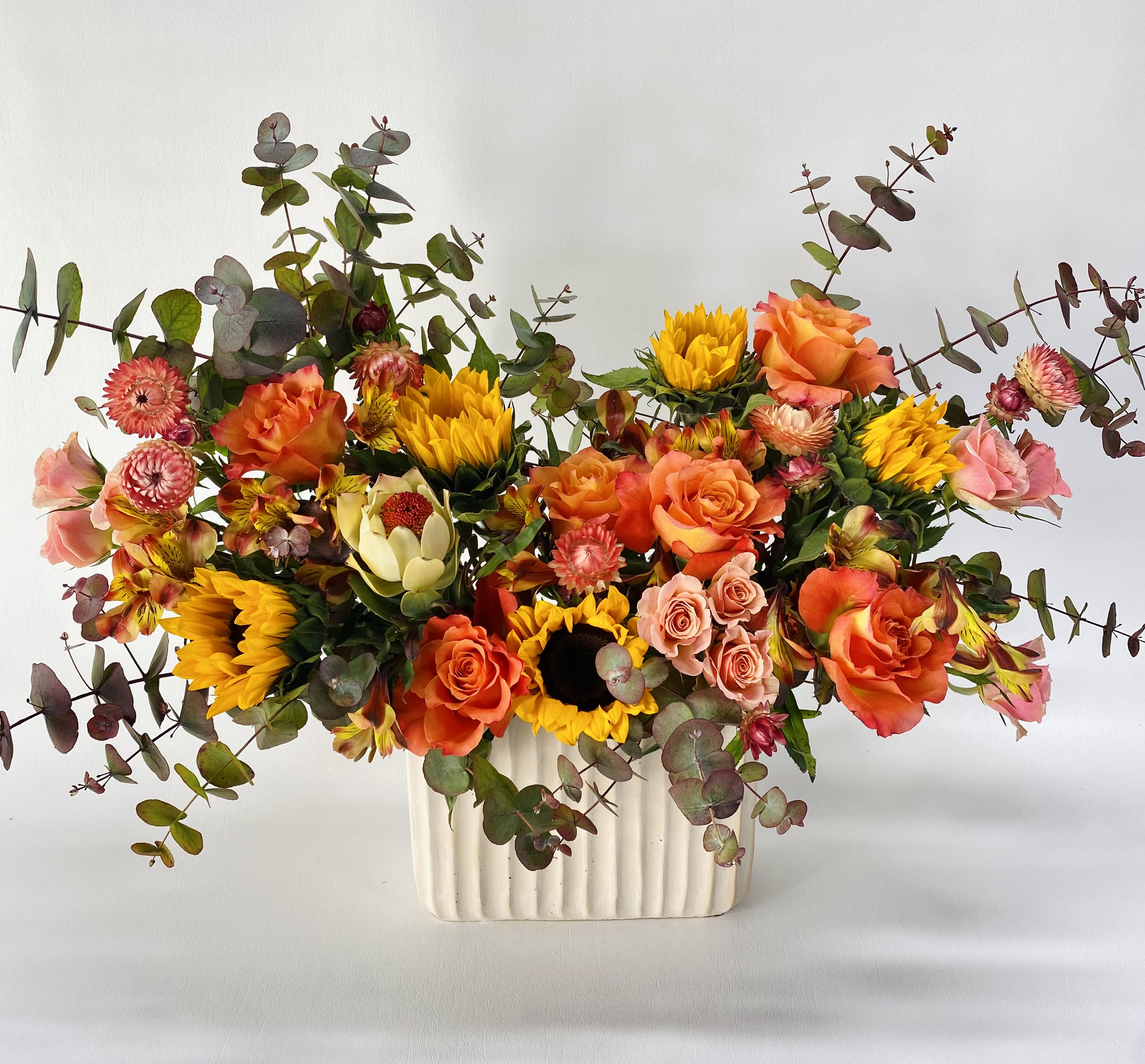 Mixed bouquet of orange roses, sunflowers, and pink blooms in a white vase