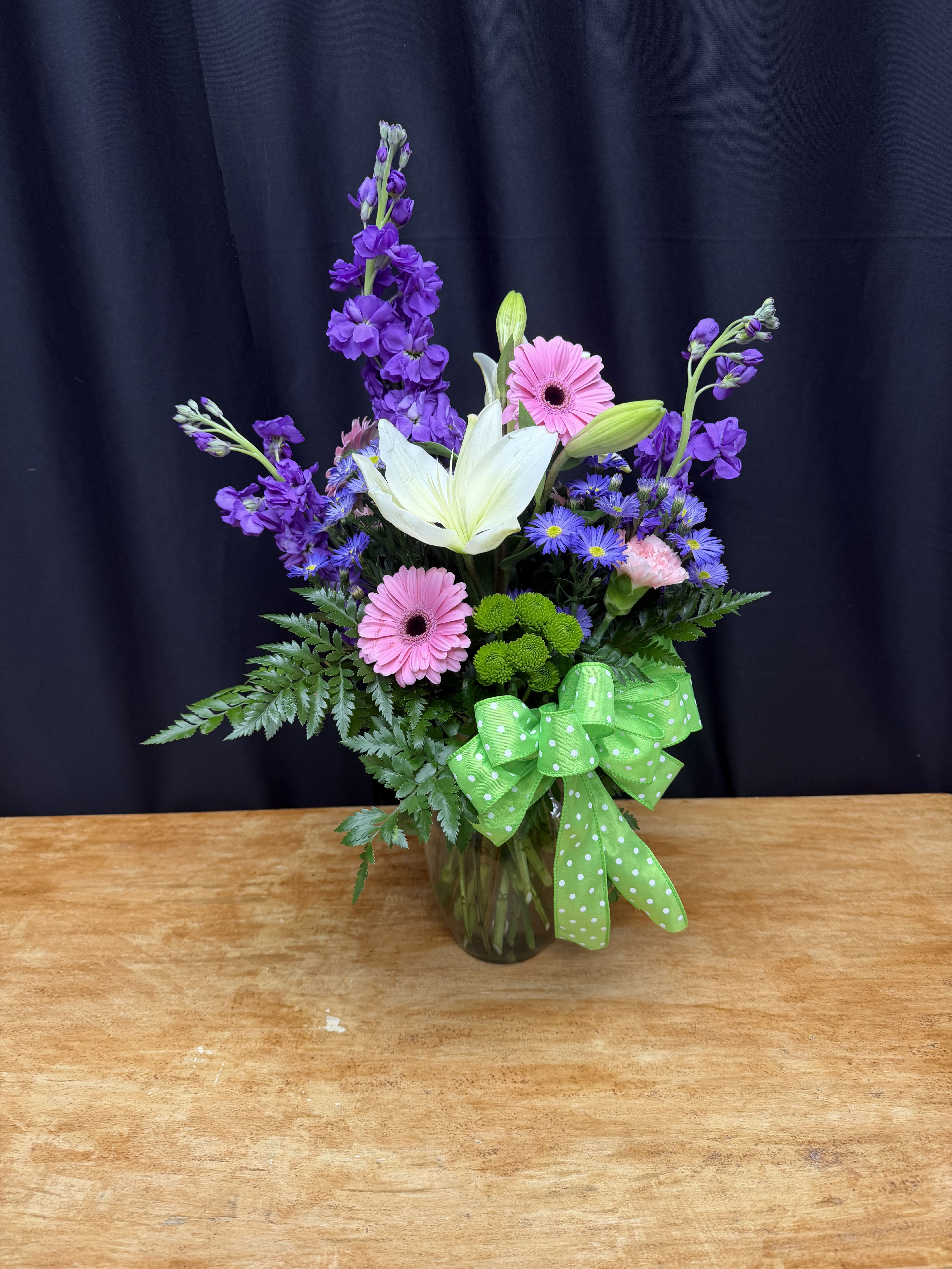 Spring Serenade - A cheerful mix of pink gerbera daisies, fragrant lavender stock, and a striking white lily creates a fresh, uplifting focal point. Accented with vibrant green button mums, delicate asters, and lush greenery, this arrangement brings a lively garden feel to