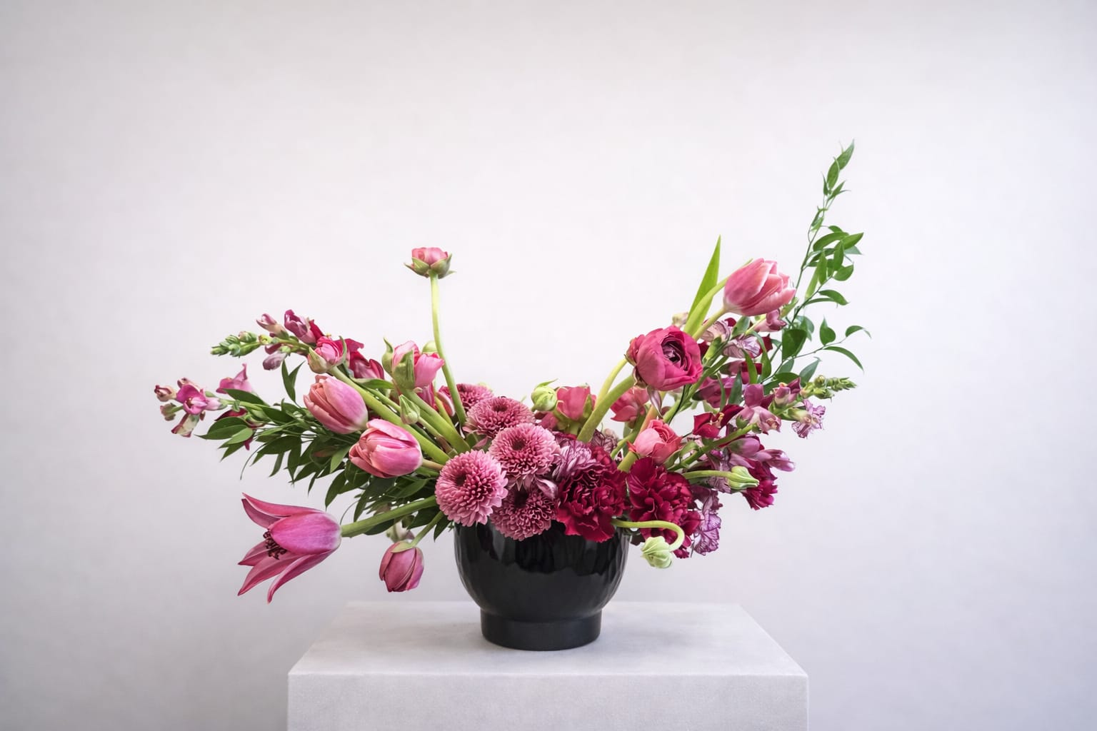 Midnight Berry Soirée - Anchored in a sleek, high-gloss obsidian vessel, the design features a lush heart of magenta chrysanthemums and velvety carnations. Sweeping outwards are graceful tulips and snapdragons that provide a playful, "just-picked" movement, while reaching greenery adds a touch of untamed elegance. It’s a bold statement piece that balances modern sophistication with romantic charm.