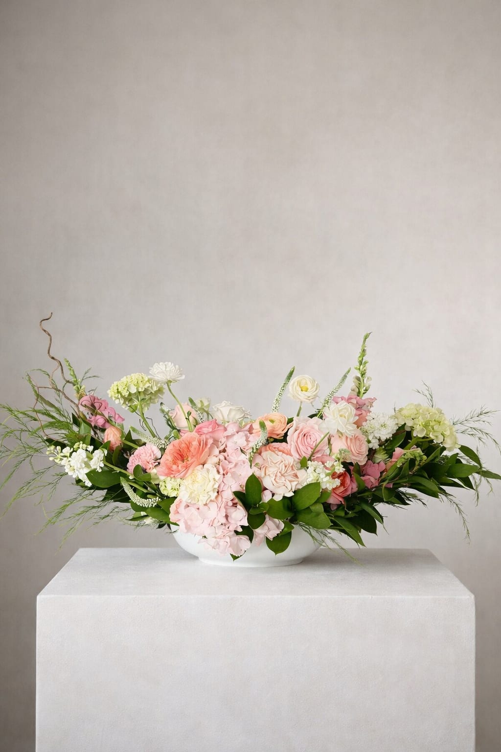 The Grand Garden Statement - Layers of blush hydrangea, peach and coral roses, and delicate ranunculus unfold organically, accented by airy greenery and sculptural branches that extend gracefully beyond the vessel.  With its low, expansive silhouette, this arrangement is designed to anchor a table or space without obstructing the view—offering a lush, immersive experience from every angle. Each bloom is thoughtfully placed to evoke the feeling of a freshly gathered garden, elevated through a modern, editorial lens.  Ideal for statement centerpieces, intimate gatherings, or moments that call for something truly unforgettable.
