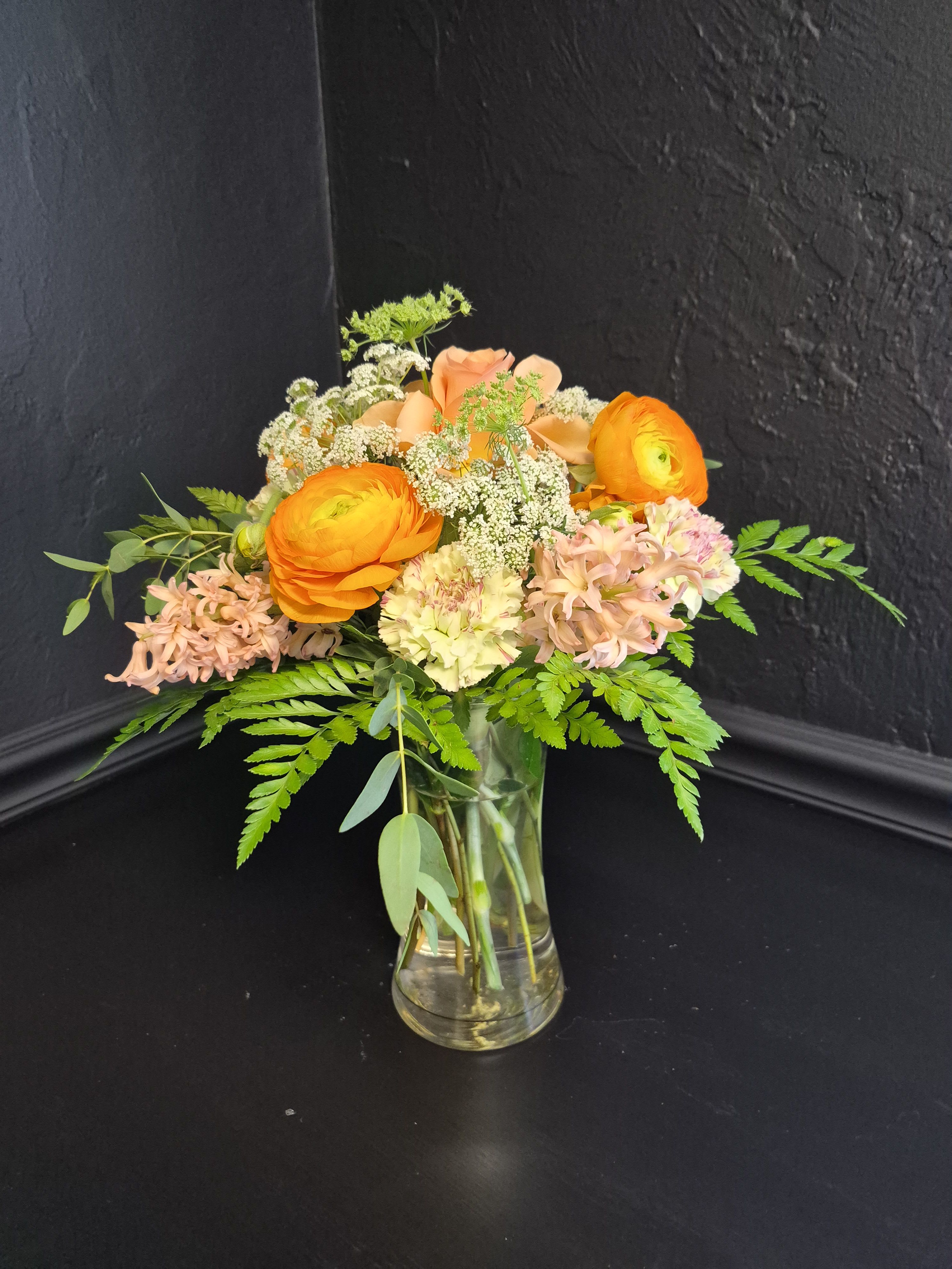 Arpicot Bloom - Bright orange ranunculus take center stage in this cheerful flower arrangement, paired with soft peach roses and pale pink hyacinths for a warm, layered birthday bouquet or spring celebration gift. Small white filler blooms and loose greenery give the design an airy, garden-style feel that makes the person you're thinking of feel instantly brightened and noticed.  The mix includes ruffled green carnations, peach-toned roses, ranunculus  blooms, hyacinths and delicate white accents arranged in a clear glass vase. Fern fronds and assorted greenery spread outward for a natural, slightly undone silhouette, offering texture and movement across the orange, peach, white, pink, and green palette.  Ideal for birthdays, thank-you gestures, or everyday brightening, this arrangement reads as both festive and approachable-the kind of bouquet that makes someone feel truly celebrated. Vase included; keep flowers in fresh water, trim stems every few days, and place away from direct heat to extend their life.