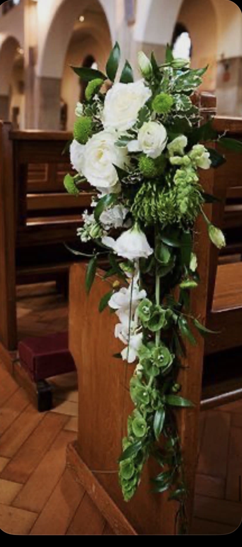 Everlasting Blooms Elegant Pew Arrangements - A creative visual expression designed for life’s many occasions, this garden-inspired floral design features elegant blooms such as roses and other similar flowers, complemented by soft green accents and textured greenery. The arrangement is styled in a graceful downward flow, creating a natural trailing effect that beautifully extends along the side of the pew.